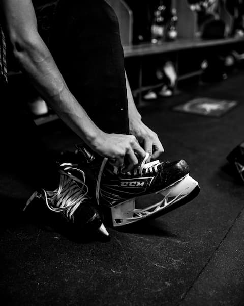 Person tying the laces on a CCM hockey skate while sitting on a bench in a locker room.