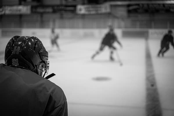 An adult attends evening skills practice, watching teammates skate on an indoor ice rink in full hockey gear.