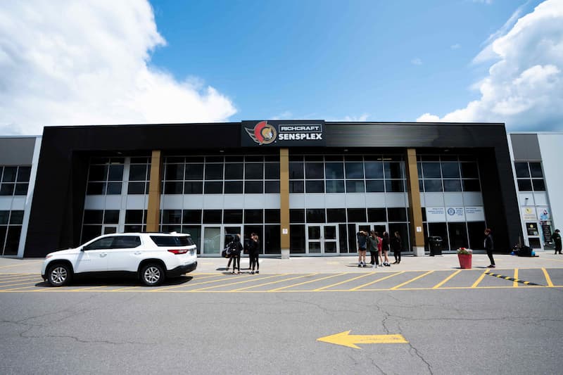Front view of a modern building labeled "Richcraft Sensplex" with people and a white SUV in the parking lot.