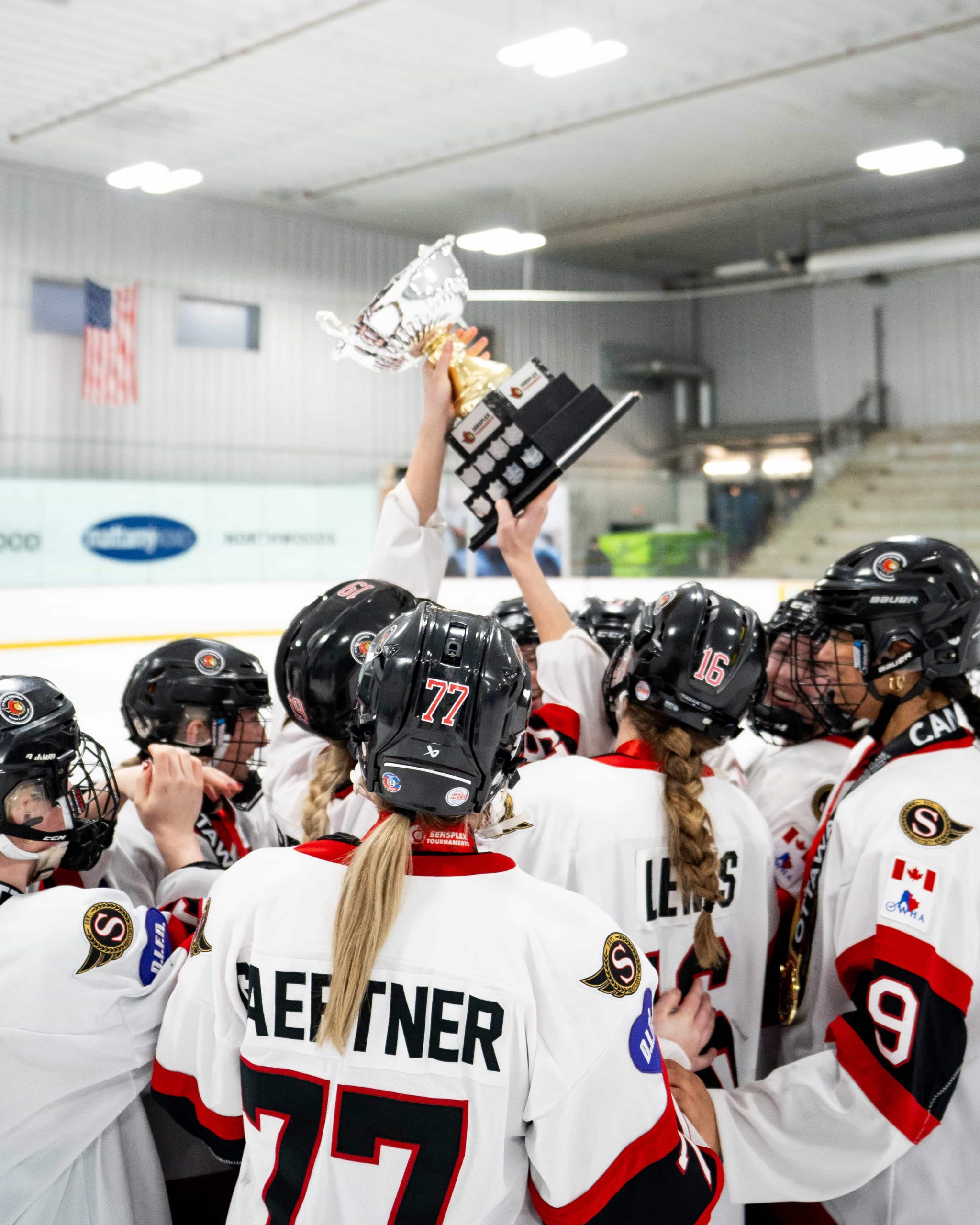 A girls’ hockey team huddles on the ice, raising a tournament trophy in celebration inside an indoor arena.
