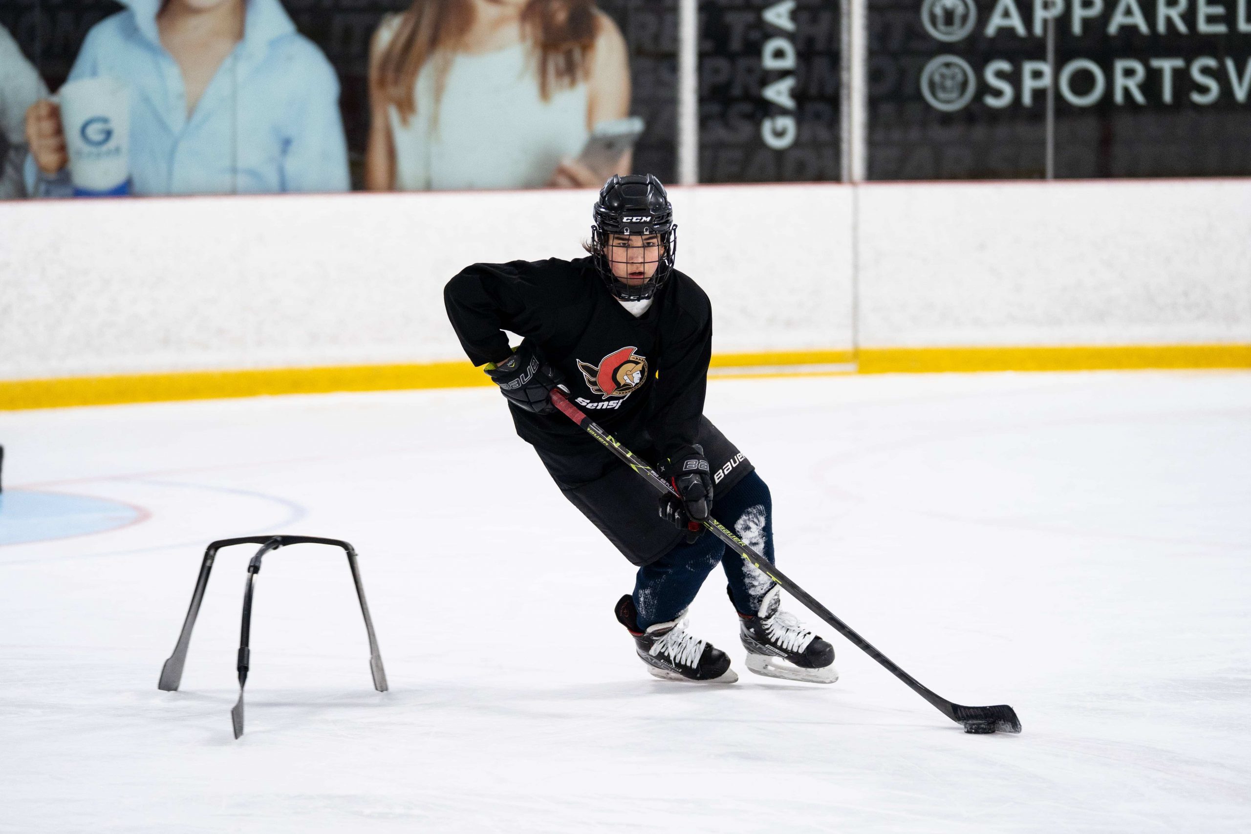 A hockey player in black gear practices skating and stickhandling on ice using a training aid.