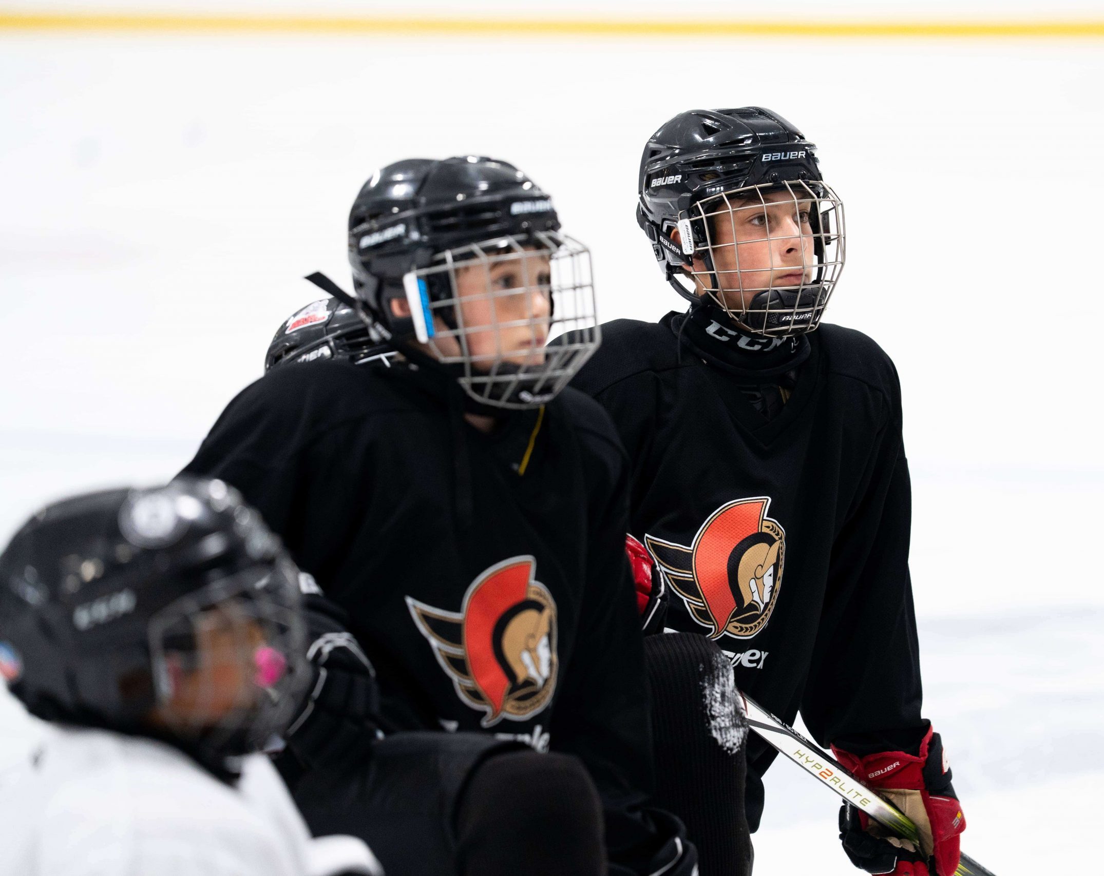 Three youth hockey players in black jerseys and helmets sit on the ice, attentively looking ahead.