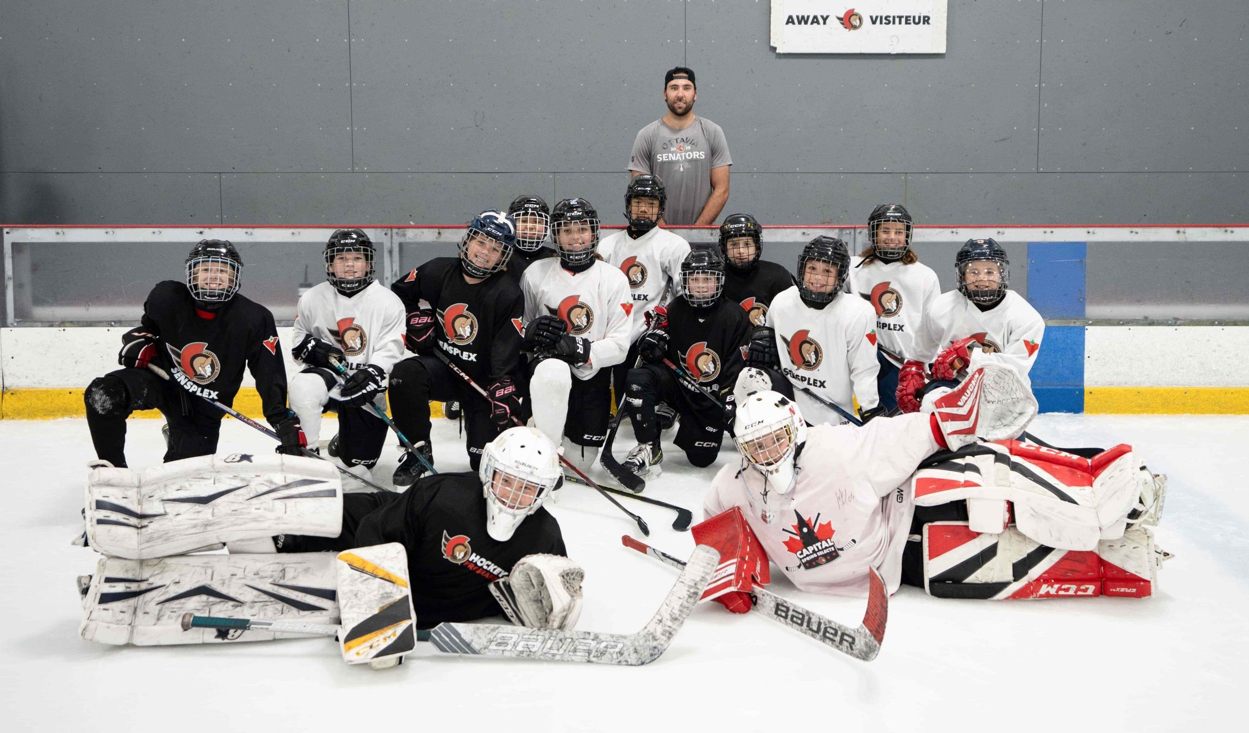 A youth hockey team in uniform poses on the ice with their coach standing behind them, in front of a rink wall.