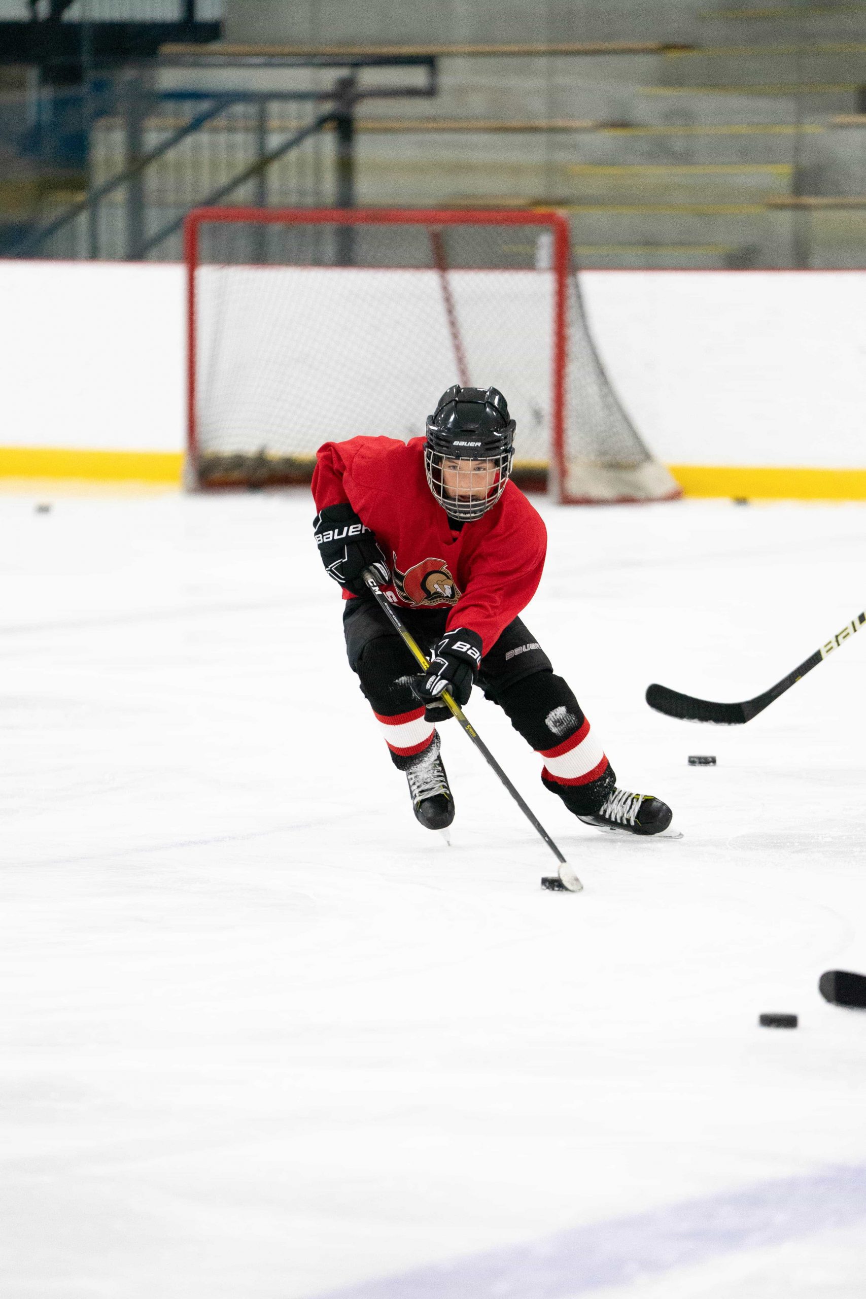 Youth hockey player in red jersey skating on ice, controlling puck with stick near the goal.