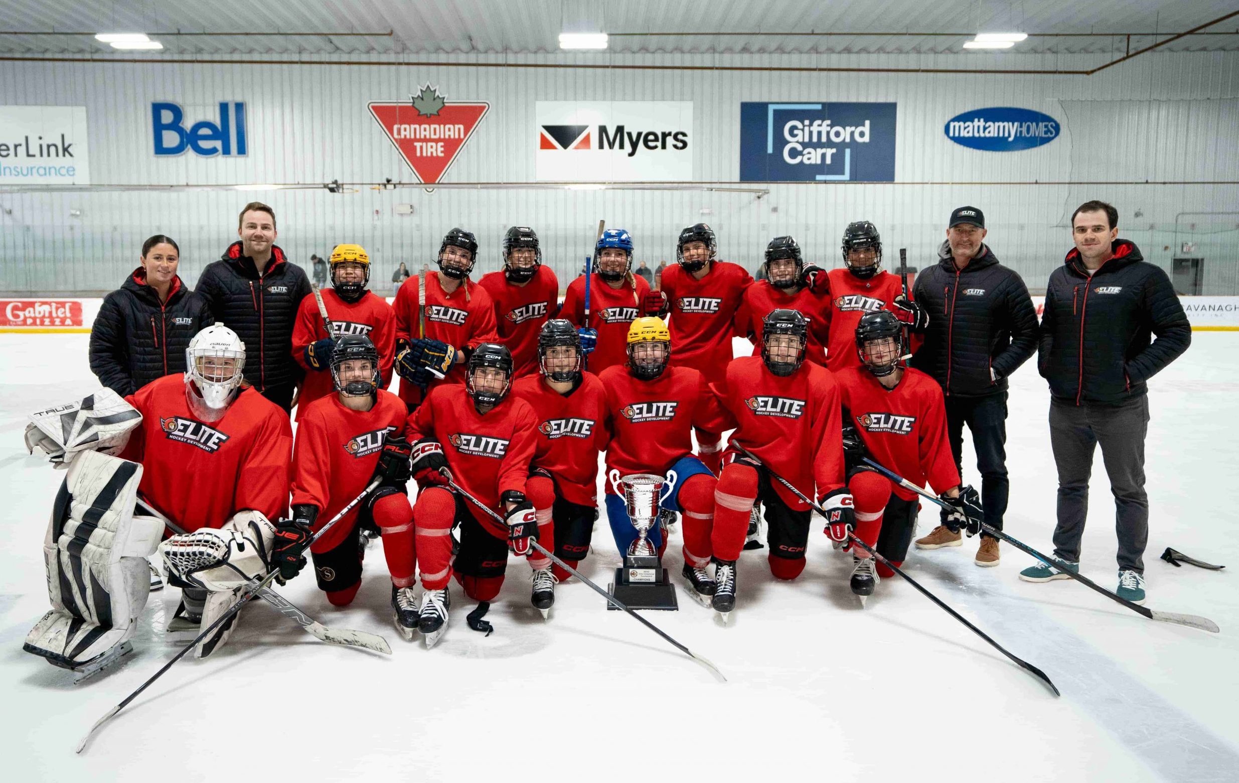 A youth hockey team in red jerseys poses on the ice with coaches and a trophy at an indoor rink.