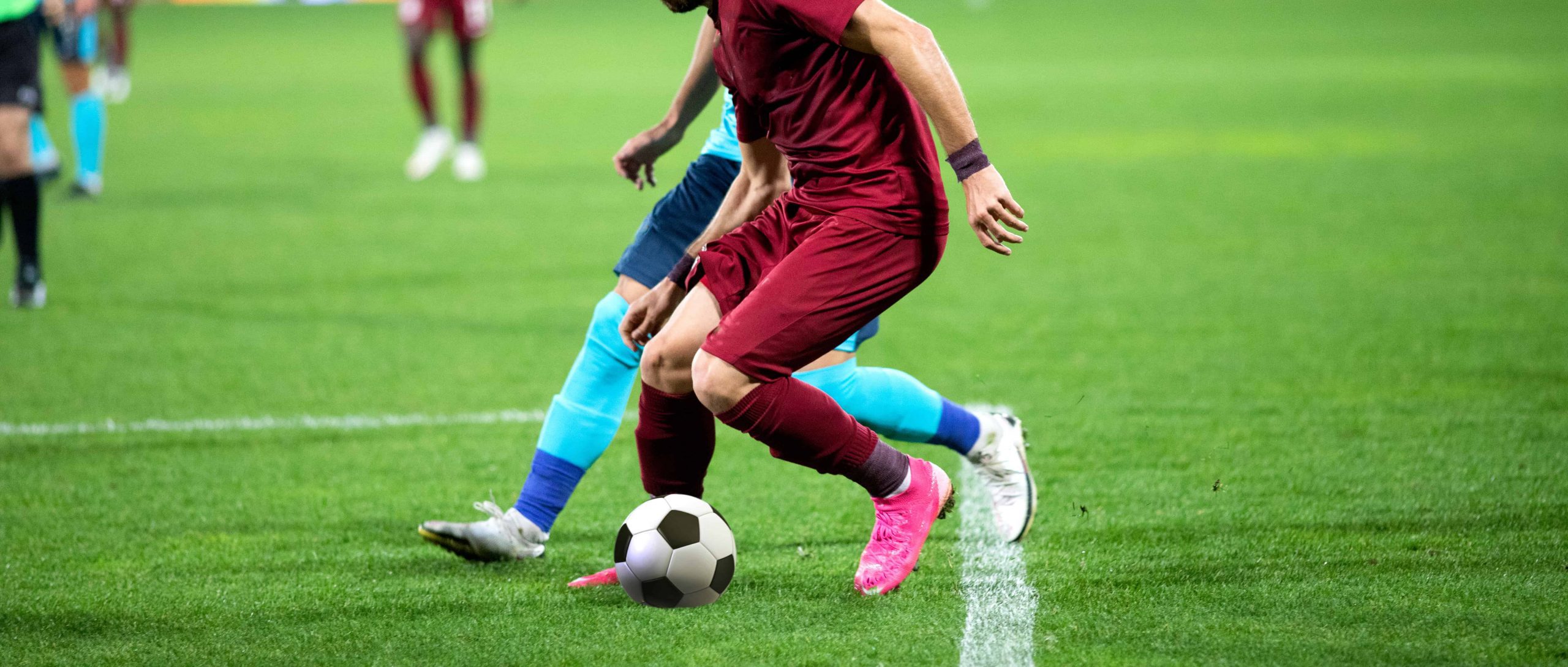Two soccer players compete for the ball on a grass field during a match, with a referee visible in the background.