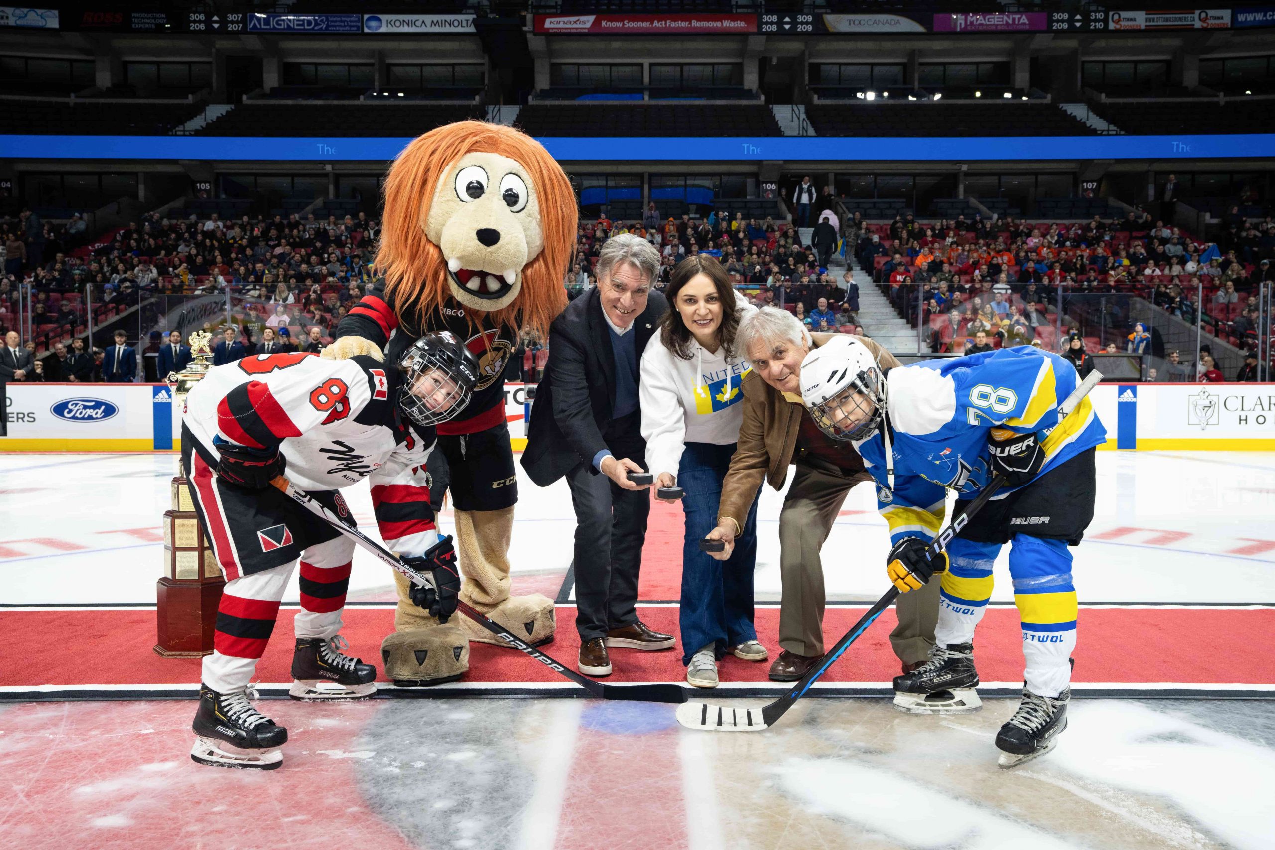Two hockey teams and three adults, joined by a mascot, pose on the ice before a face-off in an arena.