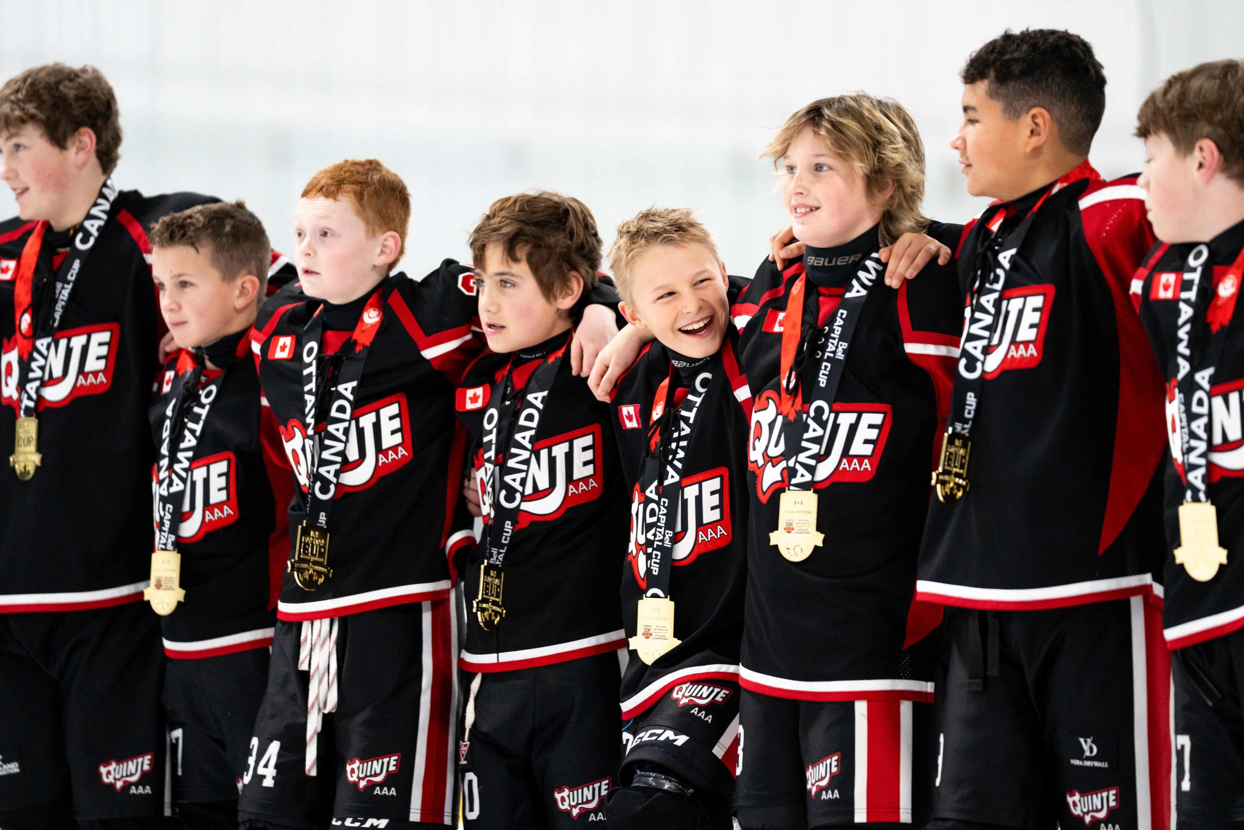 A group of young hockey players in matching black jerseys stand in a row wearing medals, with arms around each other.