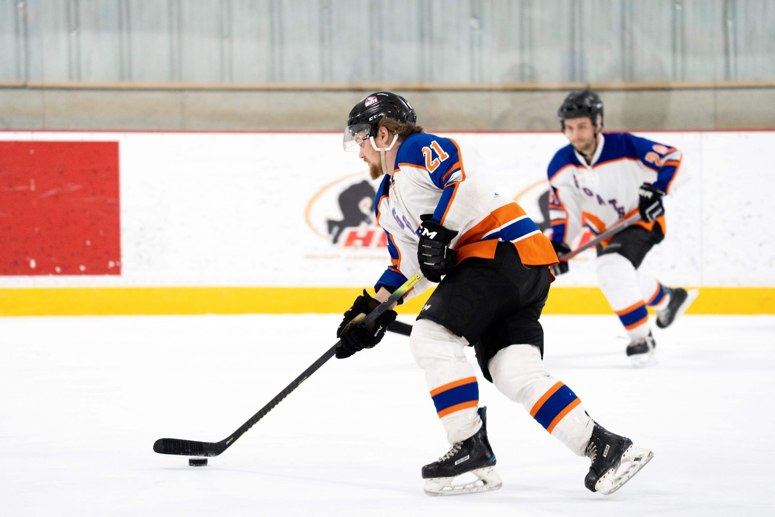 Two ice hockey players in white and blue uniforms practice adult evening skills, one handling the puck with his stick.