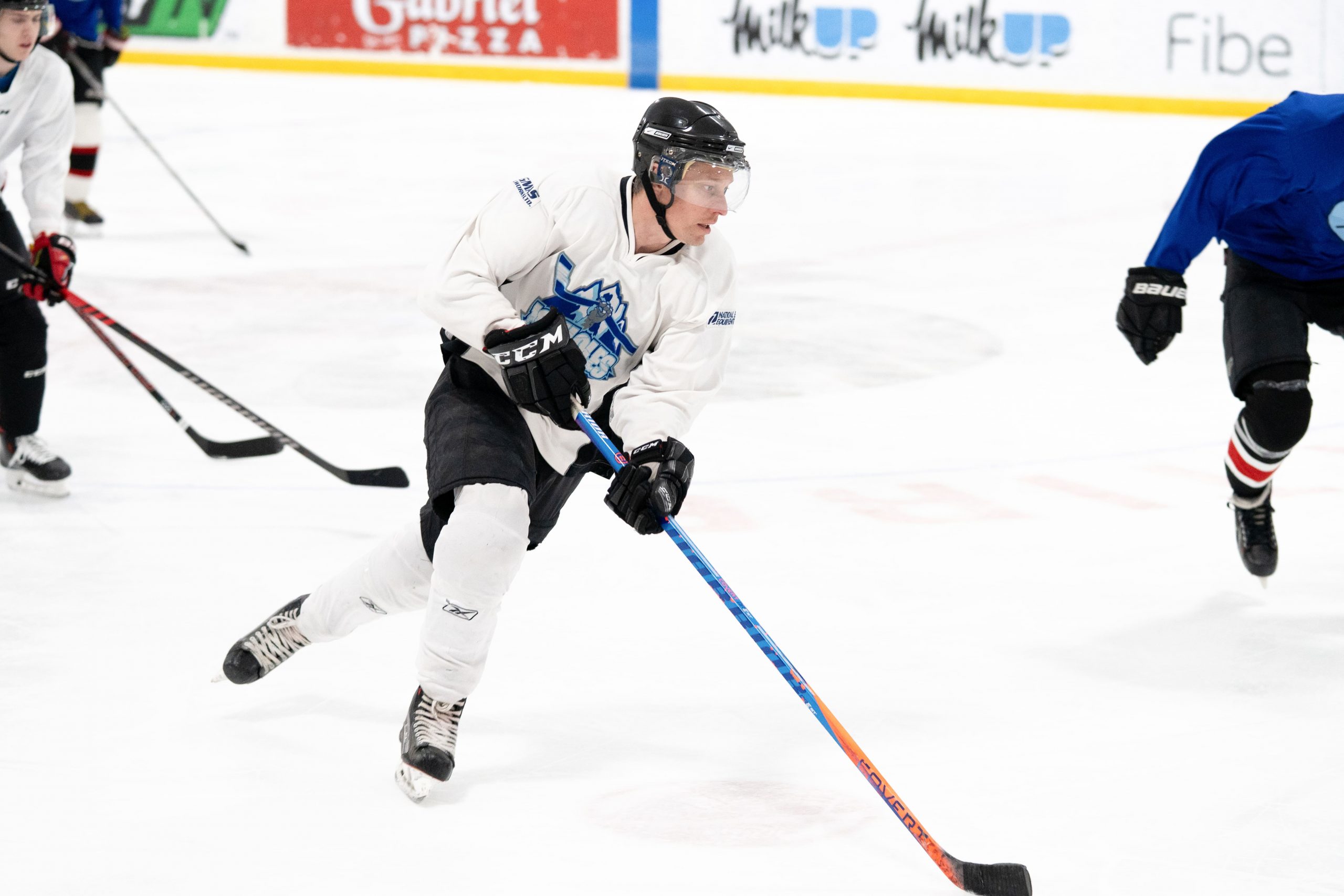 Adult evening skills on display as an ice hockey player in a white jersey skates with the puck, others in the background.