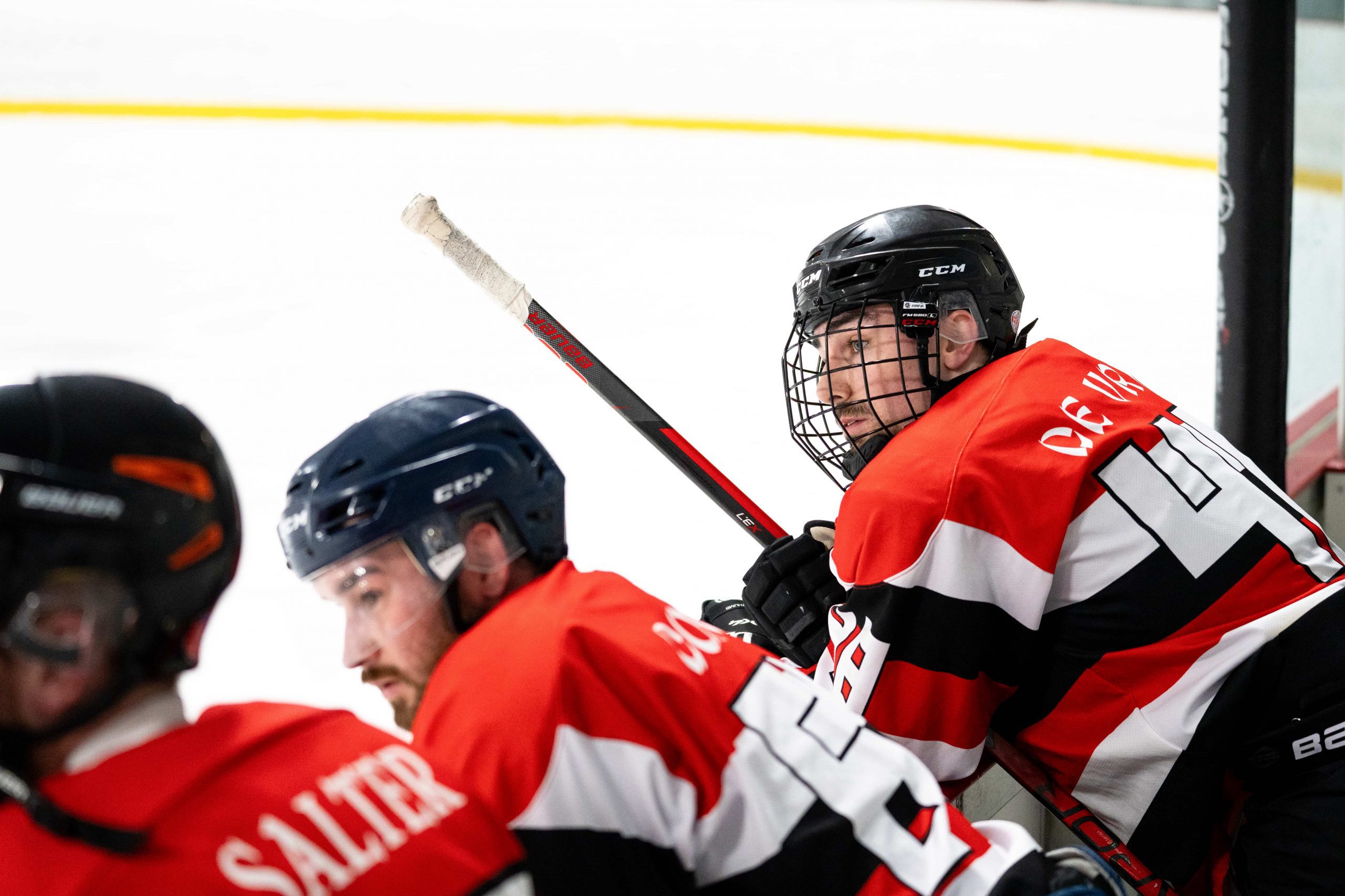 Three adult evening skills ice hockey players in red, black, and white uniforms sit on the bench during a game.