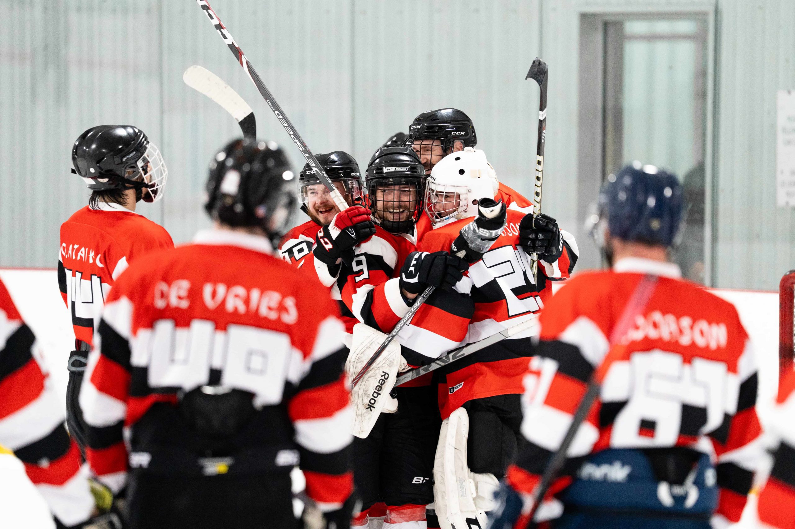 A group of adult evening skills hockey players in red and black uniforms celebrate on the ice as teammates skate toward them.