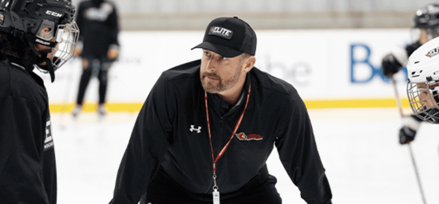 Hockey coach in black sportswear and cap talks to players on the ice during practice.