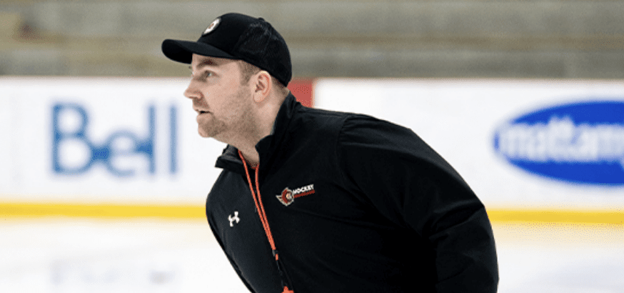 A hockey coach in black gear and a cap stands on the ice rink, with advertisements visible in the background.