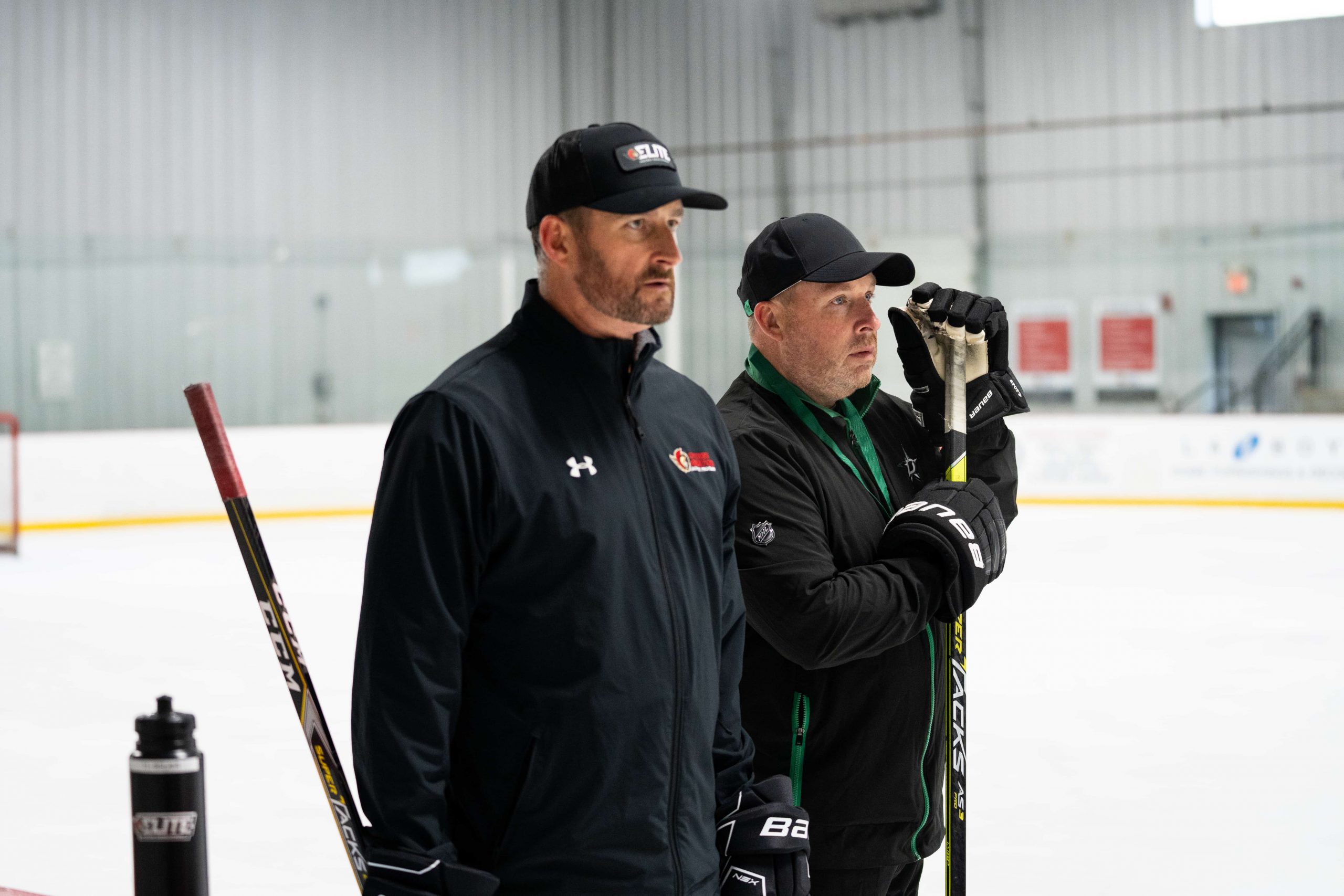 Two men in jackets and hats stand on an indoor ice rink holding hockey sticks, observing the rink.