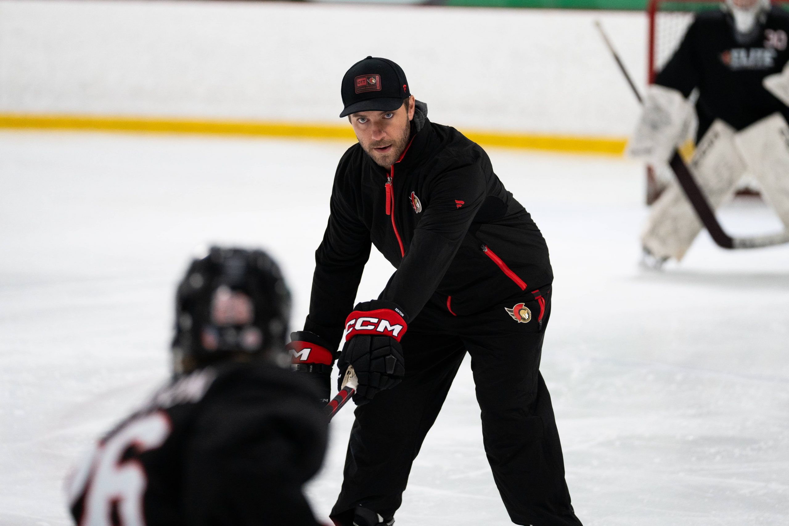 A hockey coach in black sportswear instructs a player on the ice during a practice session.