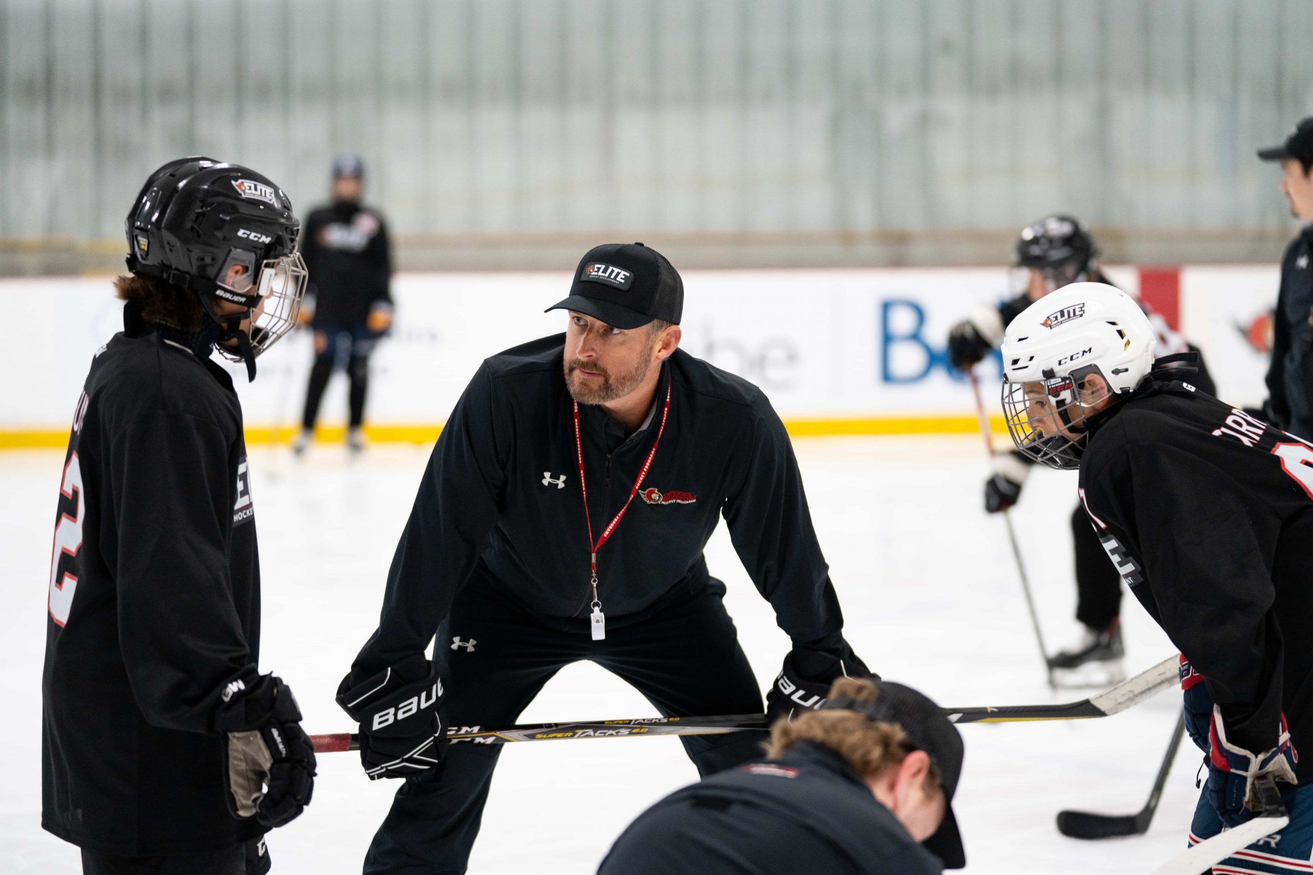A hockey coach talks to players on the ice during practice, all wearing black uniforms and helmets.