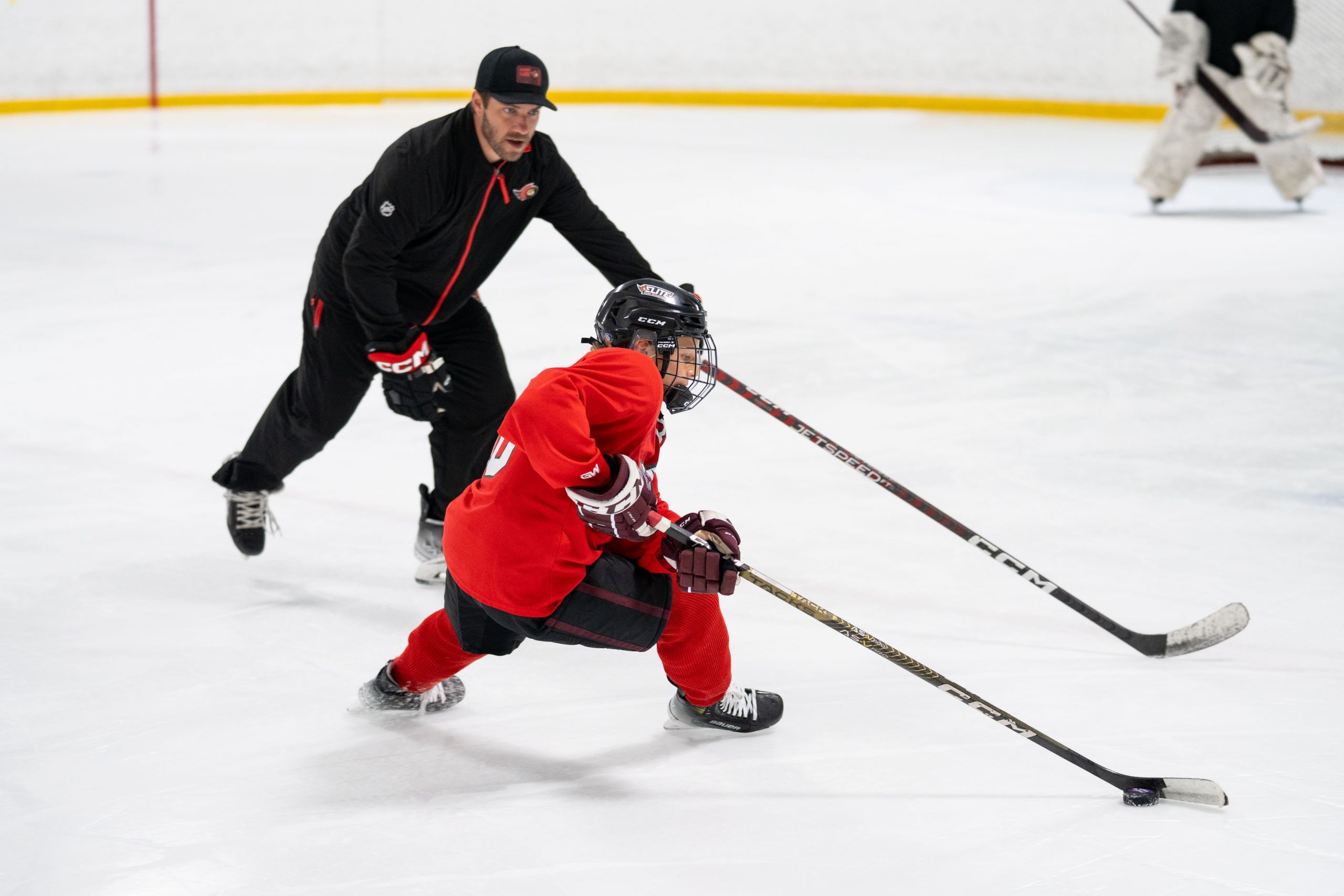 A youth hockey player in red practices skating and stickhandling on the ice with a coach nearby.