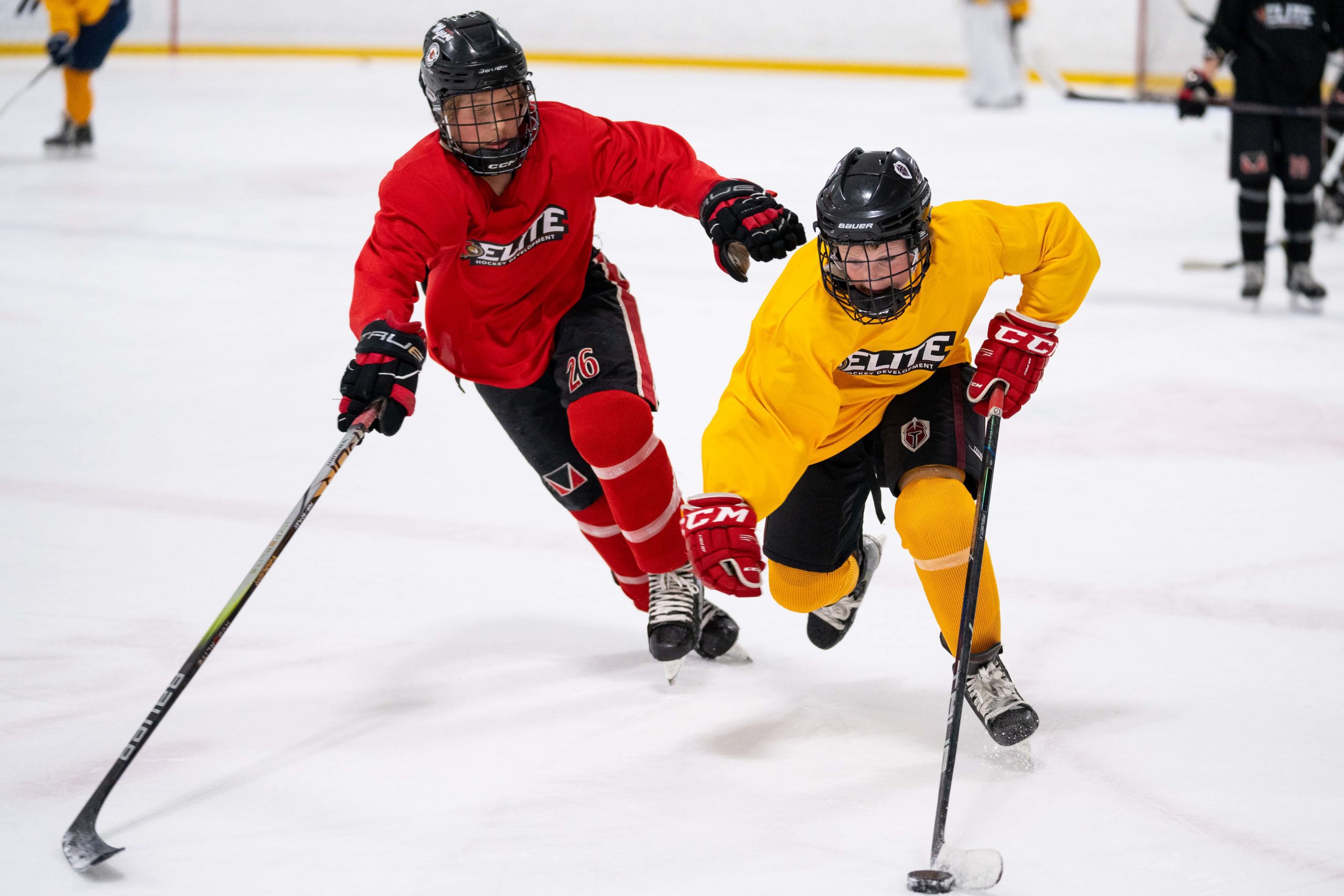 Two youth hockey players, one in red and one in yellow, compete for the puck on an indoor ice rink.