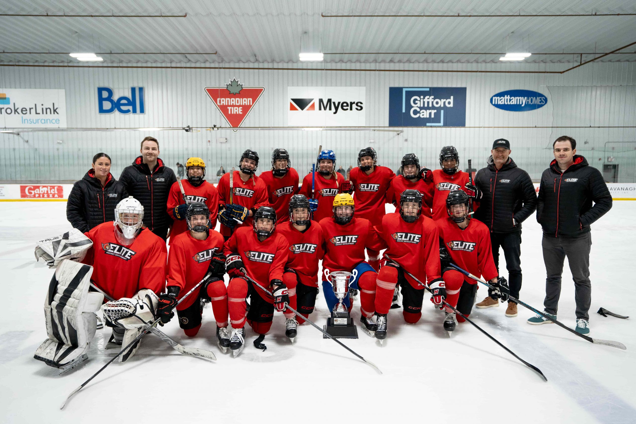 A hockey team in red jerseys poses on the ice with coaches and a trophy in an indoor rink.