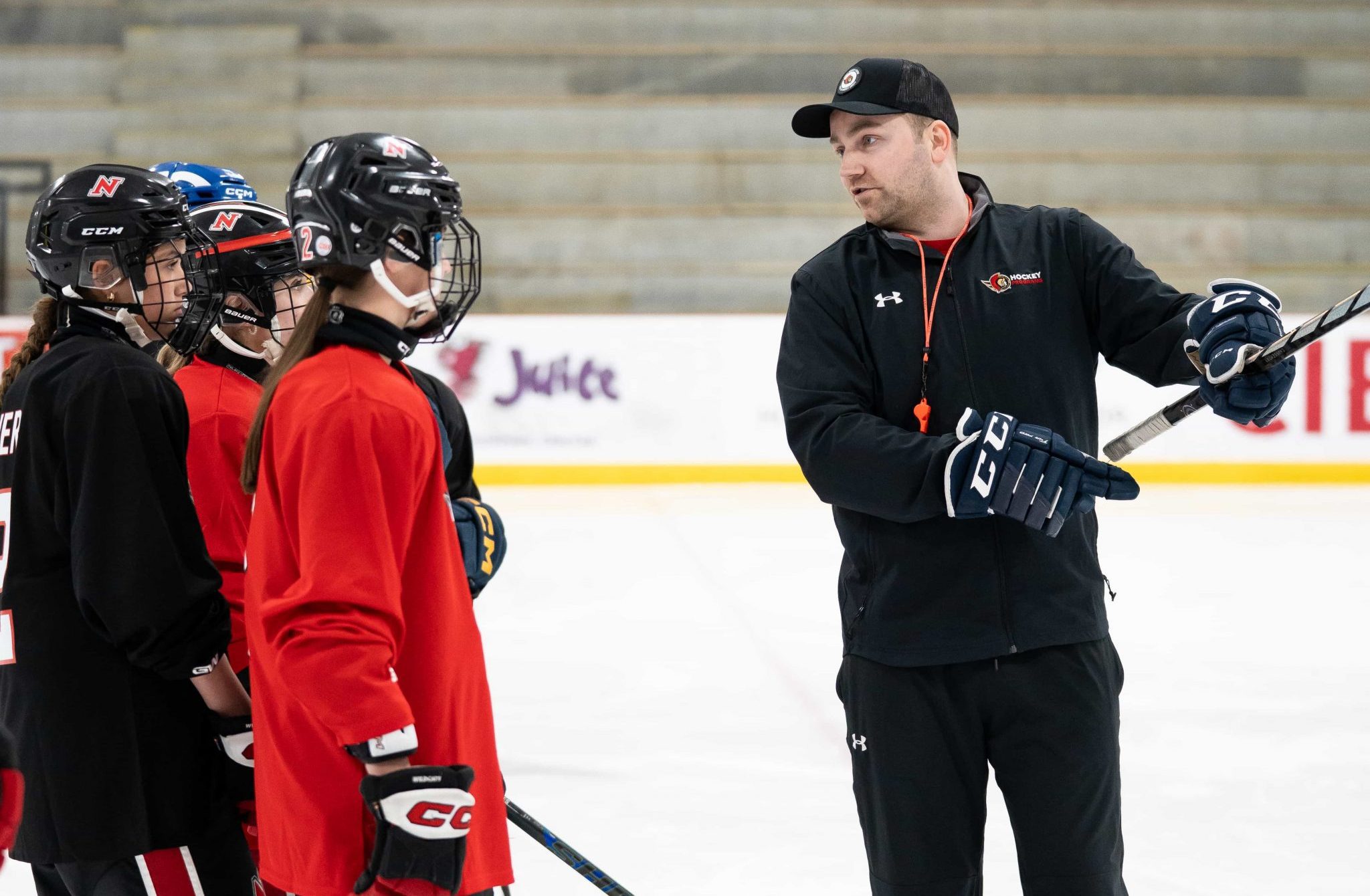 A hockey coach gives instructions to players in red and black uniforms on an indoor ice rink.