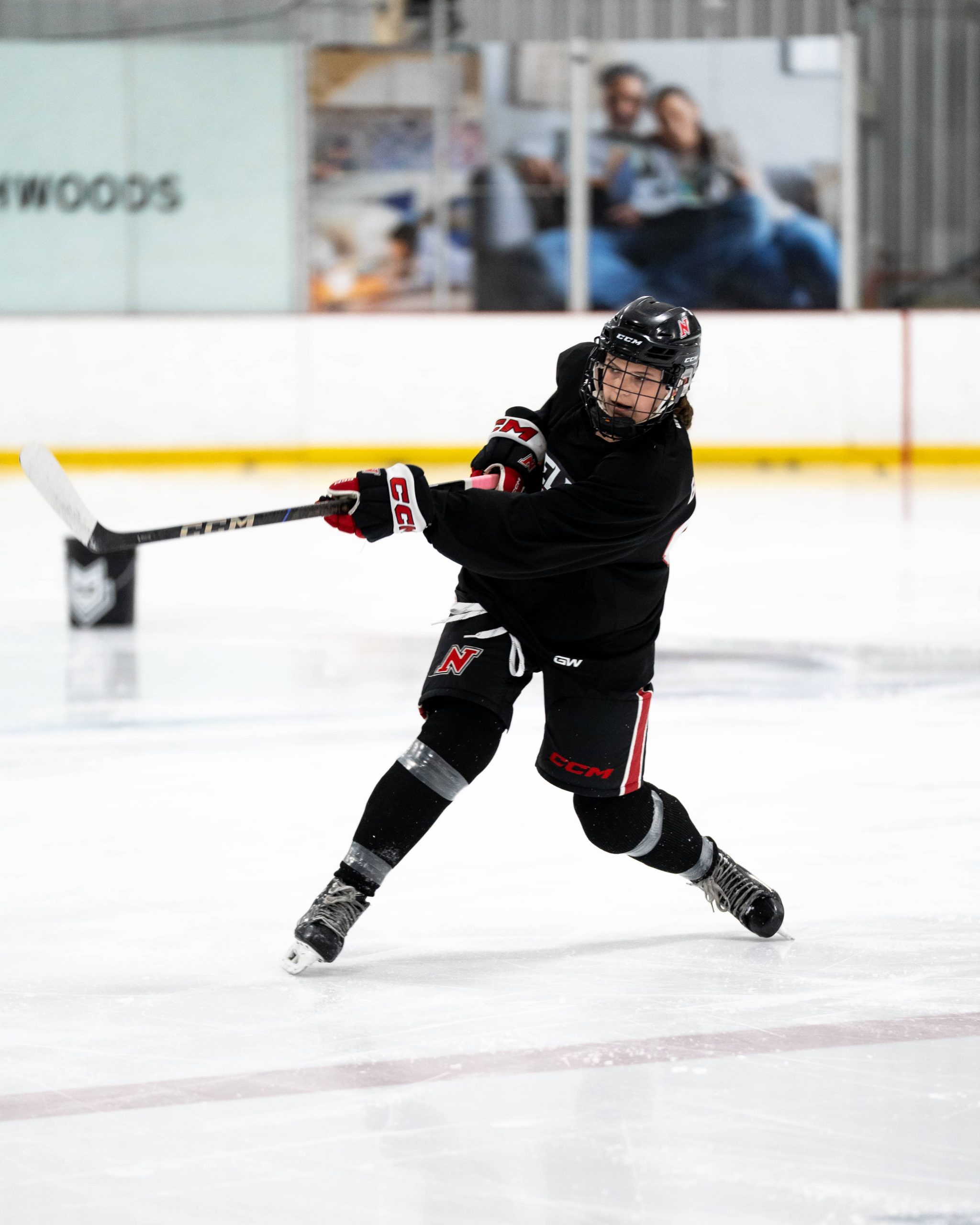 Hockey player in black gear taking a slapshot on an indoor ice rink.