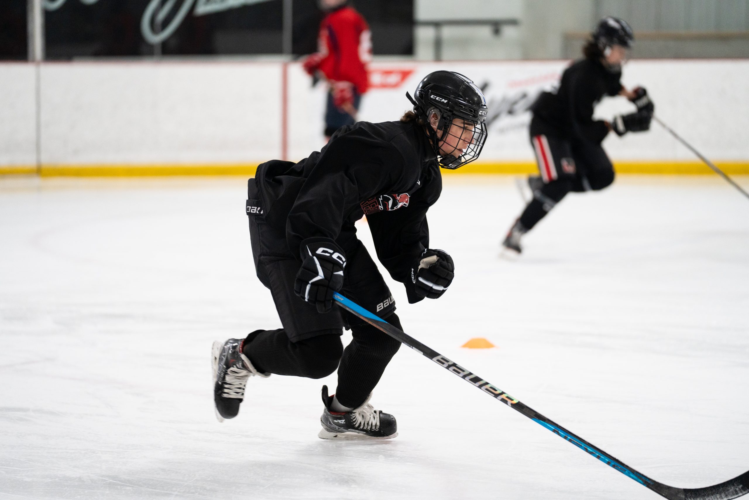 Ice hockey player in black gear skating on the rink, holding a hockey stick, with others in the background.