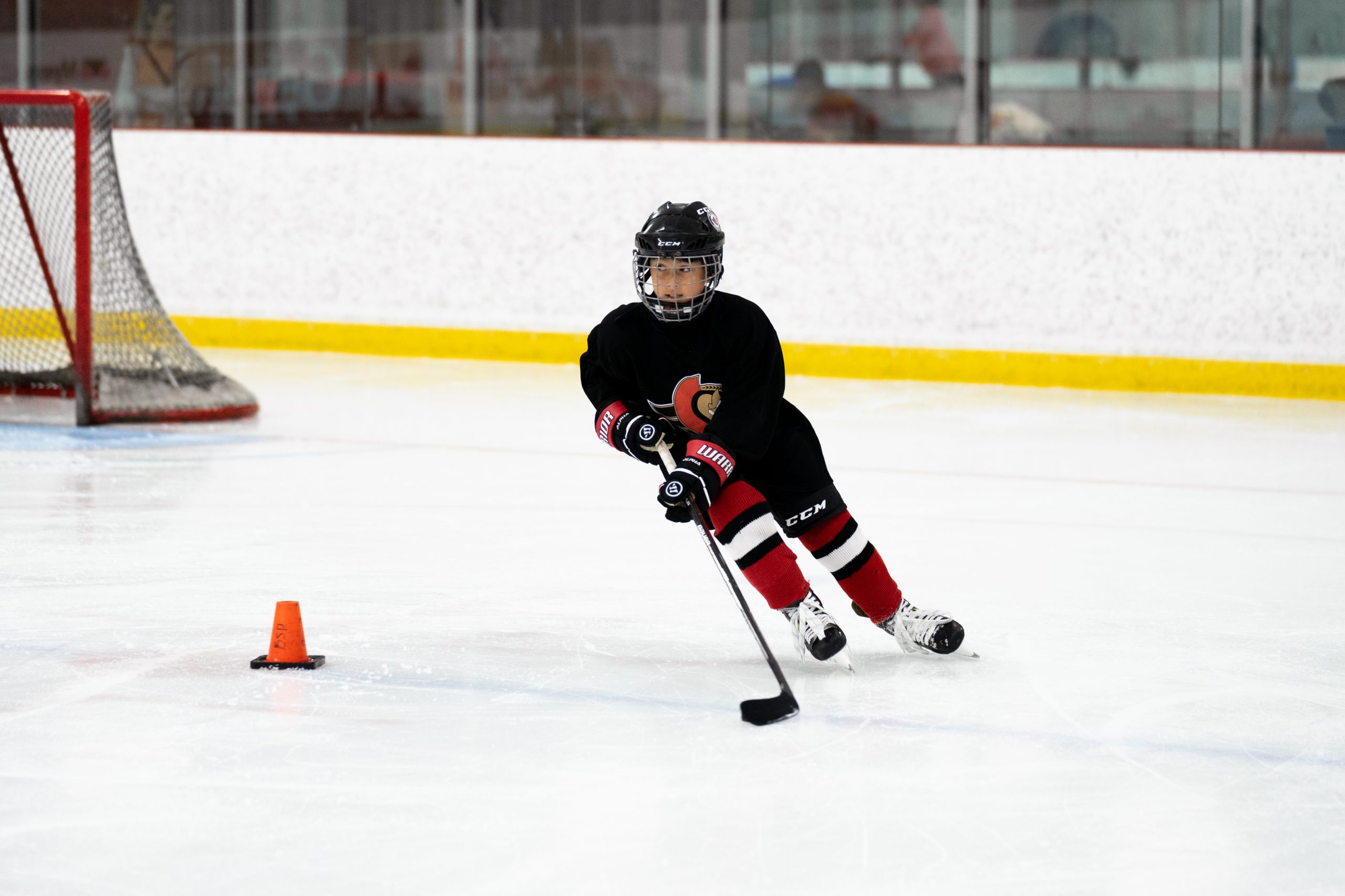 A child in hockey gear skates on an ice rink, handling a puck near an orange cone with a goal in the background.