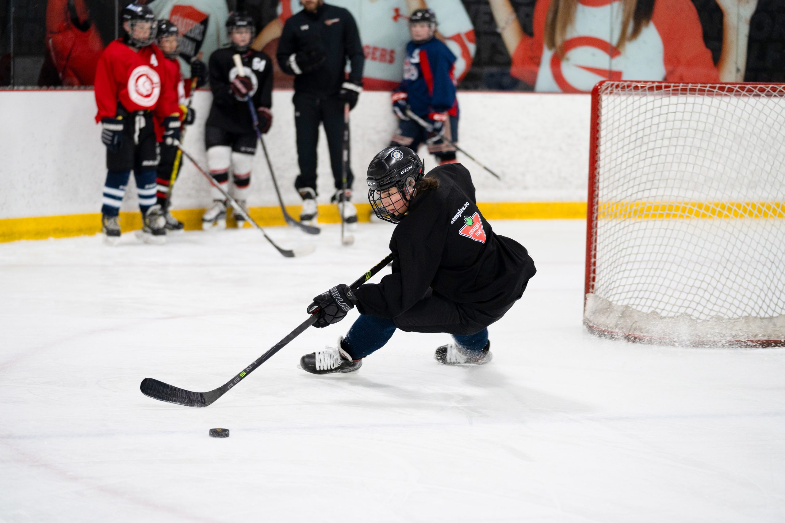 A hockey player bends low to the ice to control the puck while others watch near the goal net.