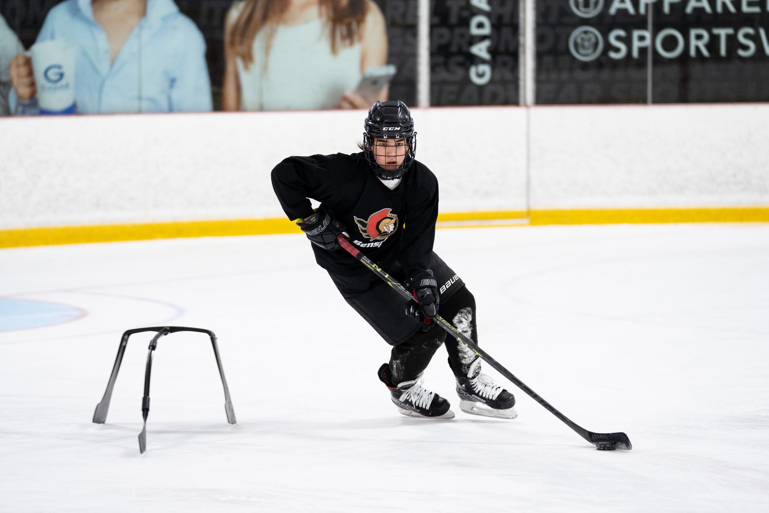 A hockey player in black gear practices skating on the ice using a metal training aid, holding a hockey stick.