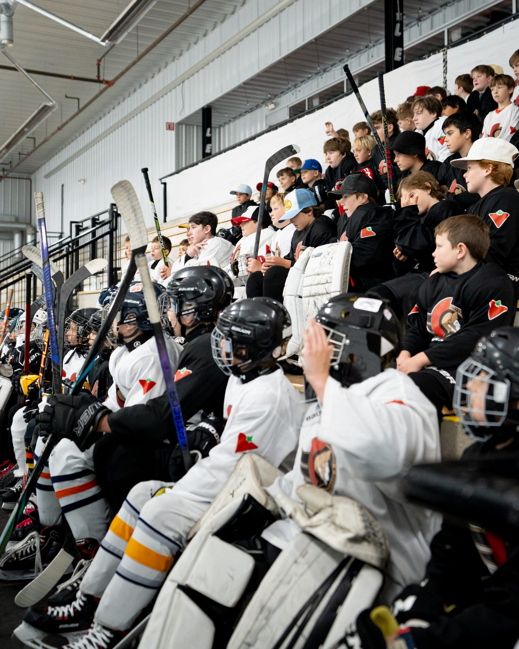 A group of young hockey players in uniform sit in bleachers, holding sticks and wearing helmets and gear.