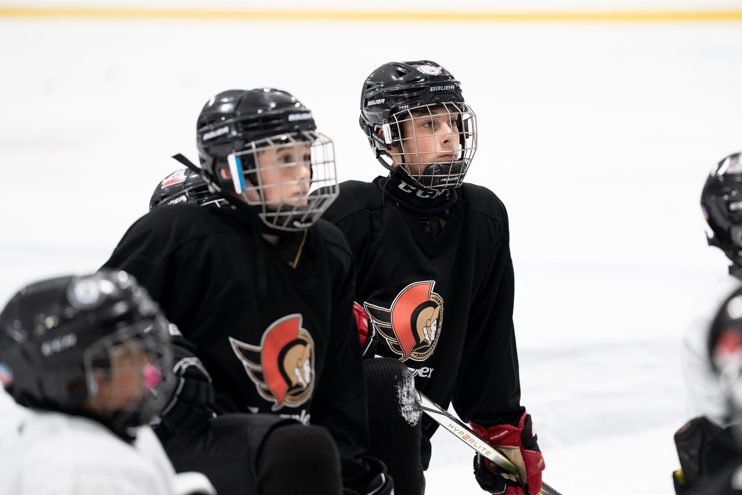Two young hockey players in black jerseys and helmets kneel on the ice, looking forward during practice.