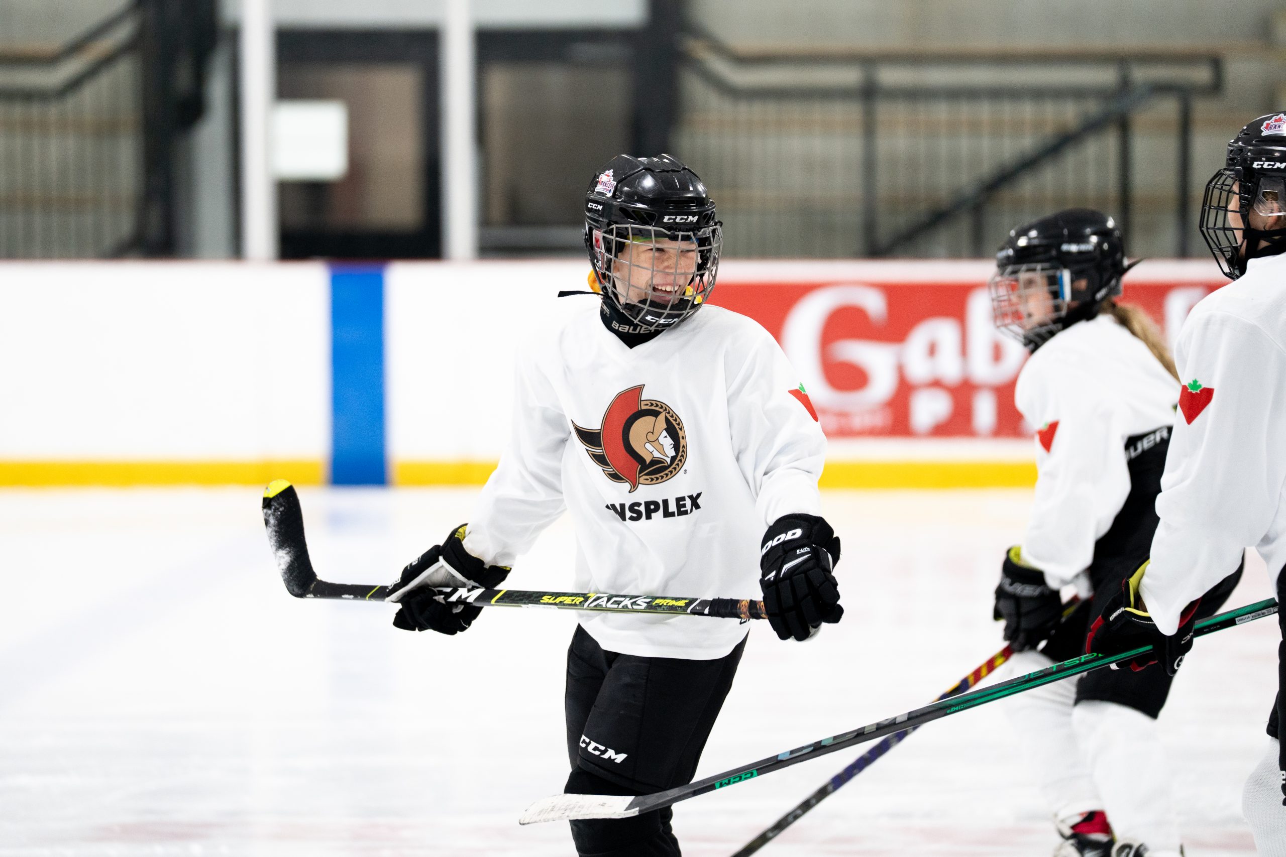 Young hockey players in white jerseys stand on an ice rink, wearing helmets and holding hockey sticks.