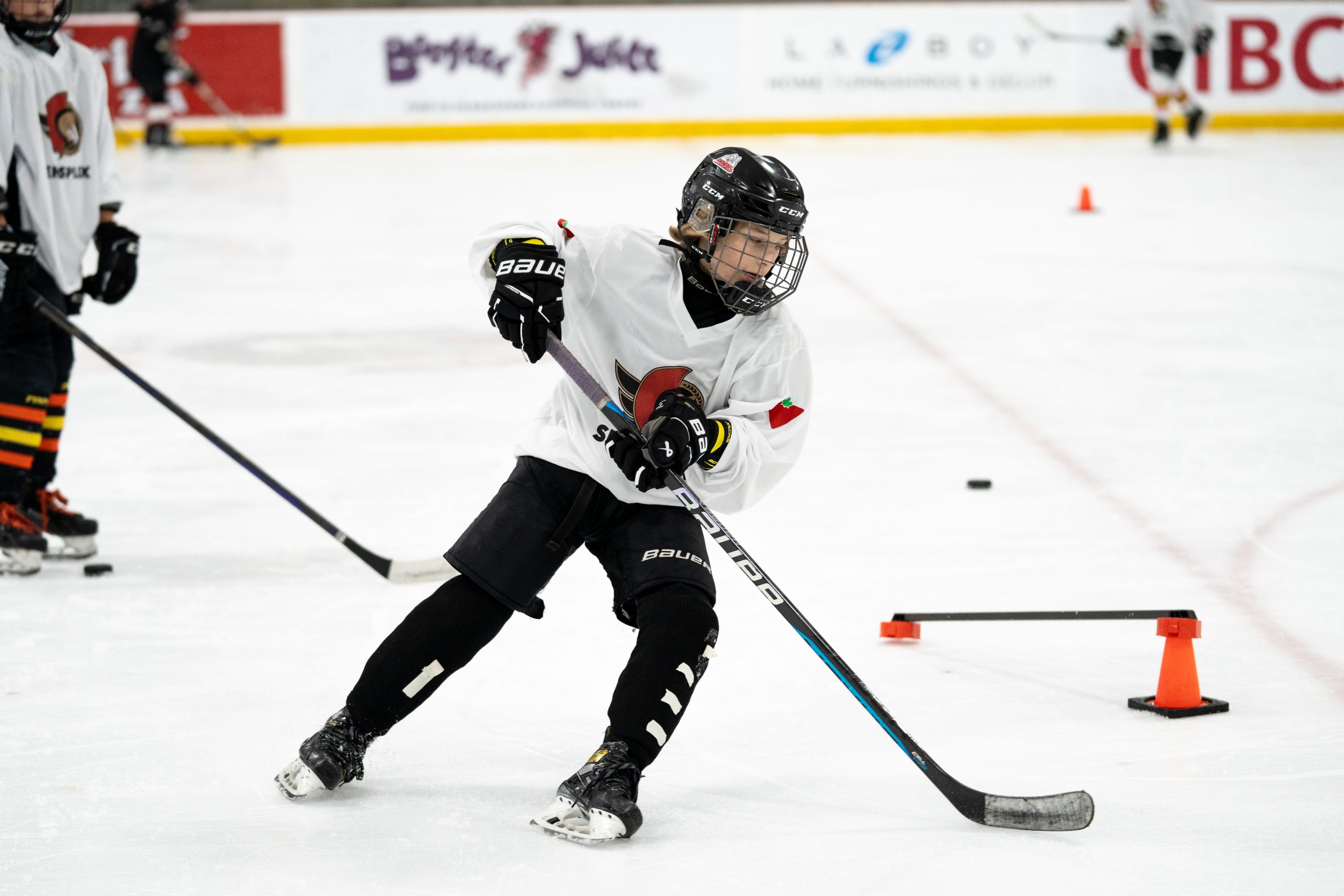 Young hockey player in white jersey practicing on ice rink, maneuvering puck around orange cones.