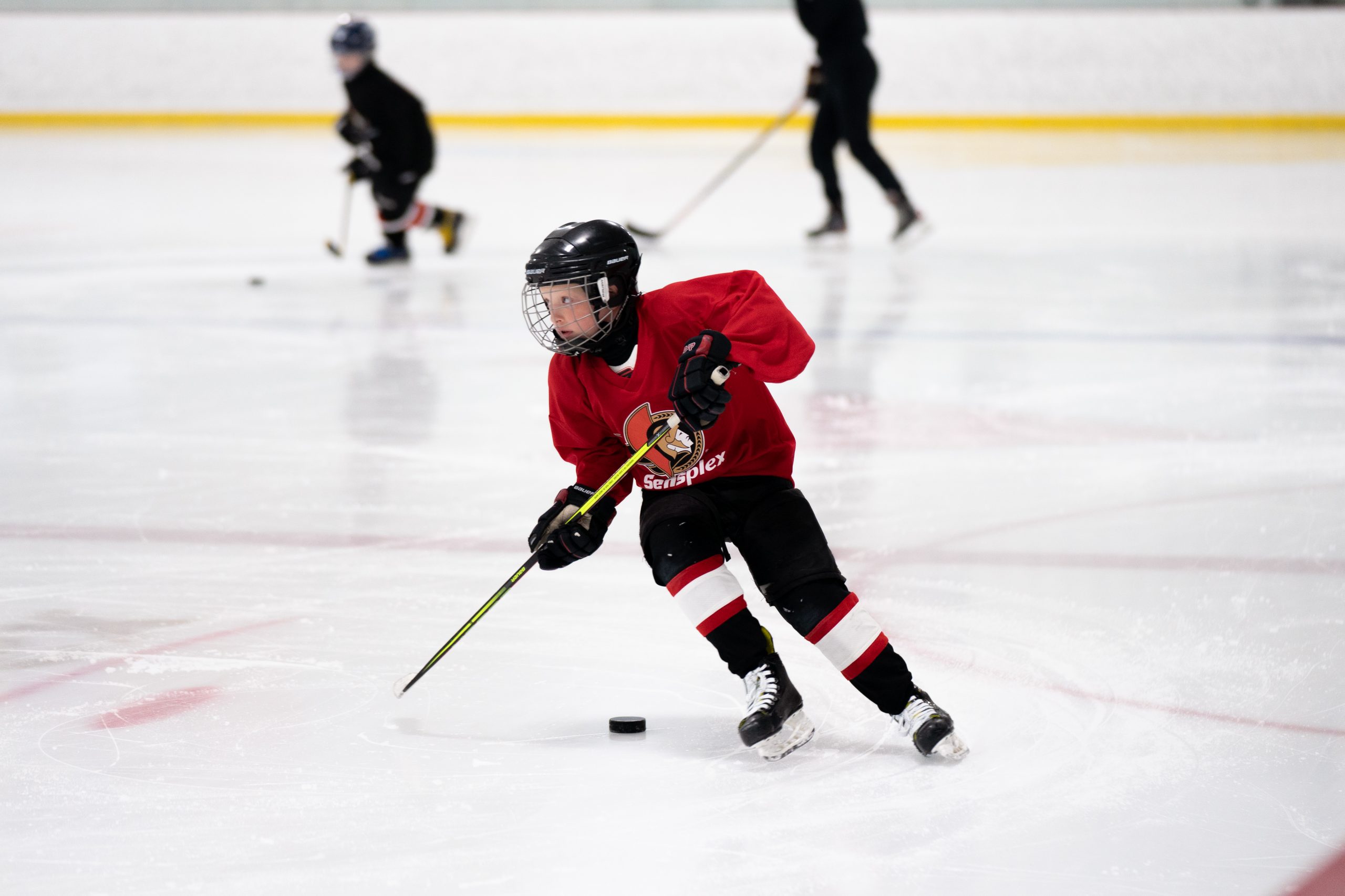 Youth hockey player in red jersey skating with a stick and puck on an ice rink, with other players in the background.