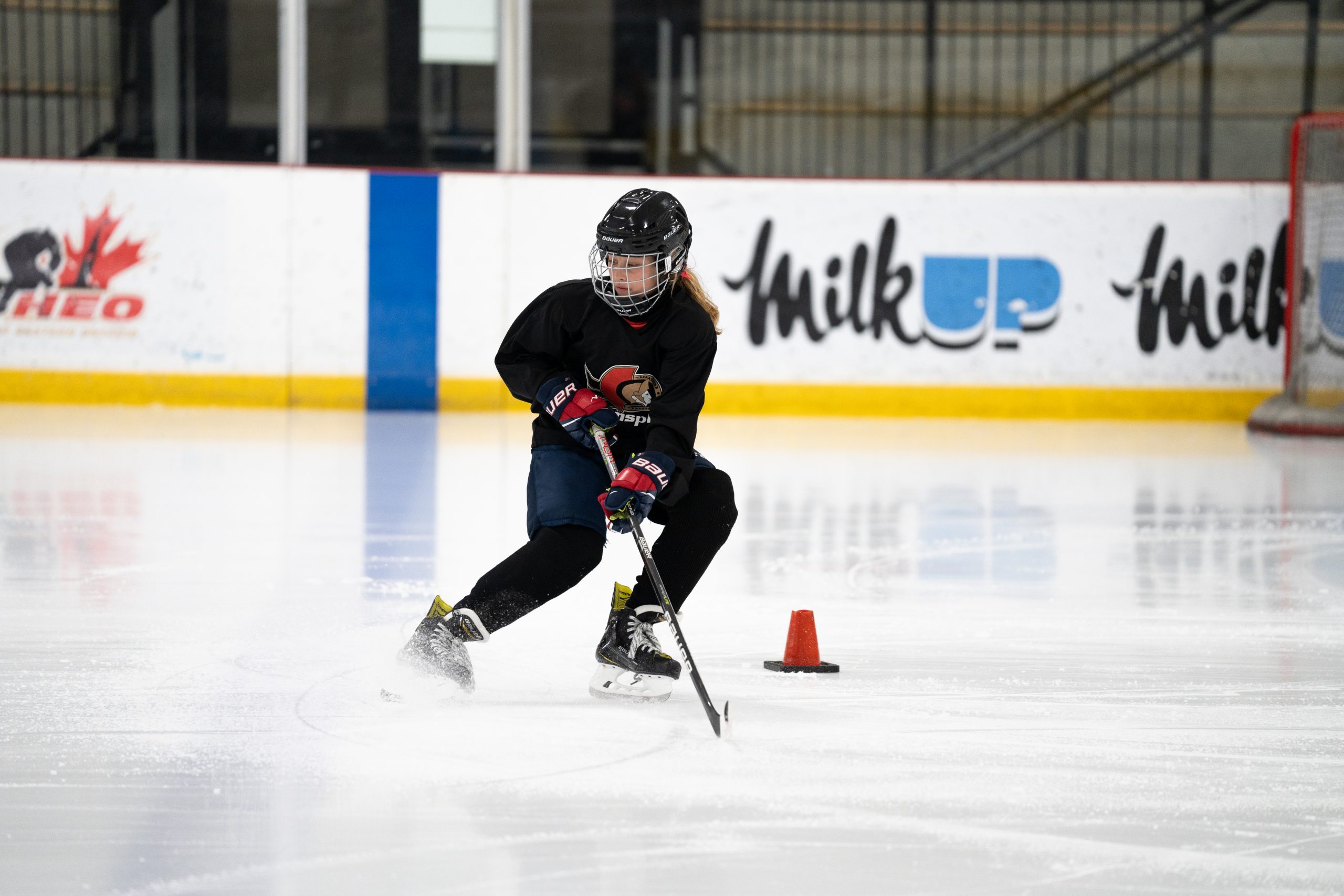 Hockey player in black jersey skating on ice with stick, maneuvering around an orange cone during practice.