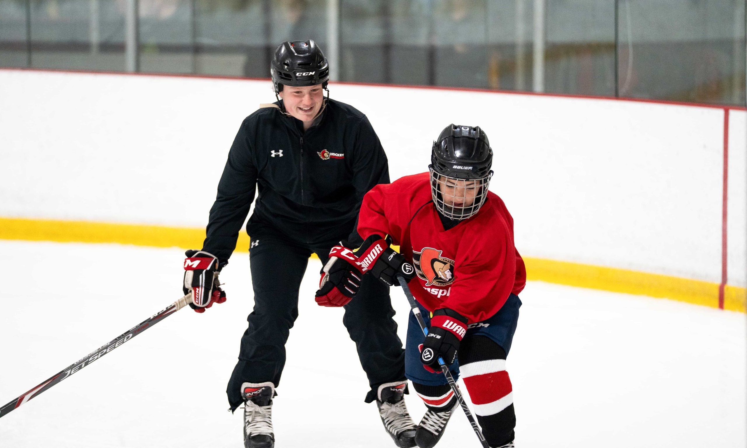 A hockey coach in black instructs a child in red gear during an on-ice drill.