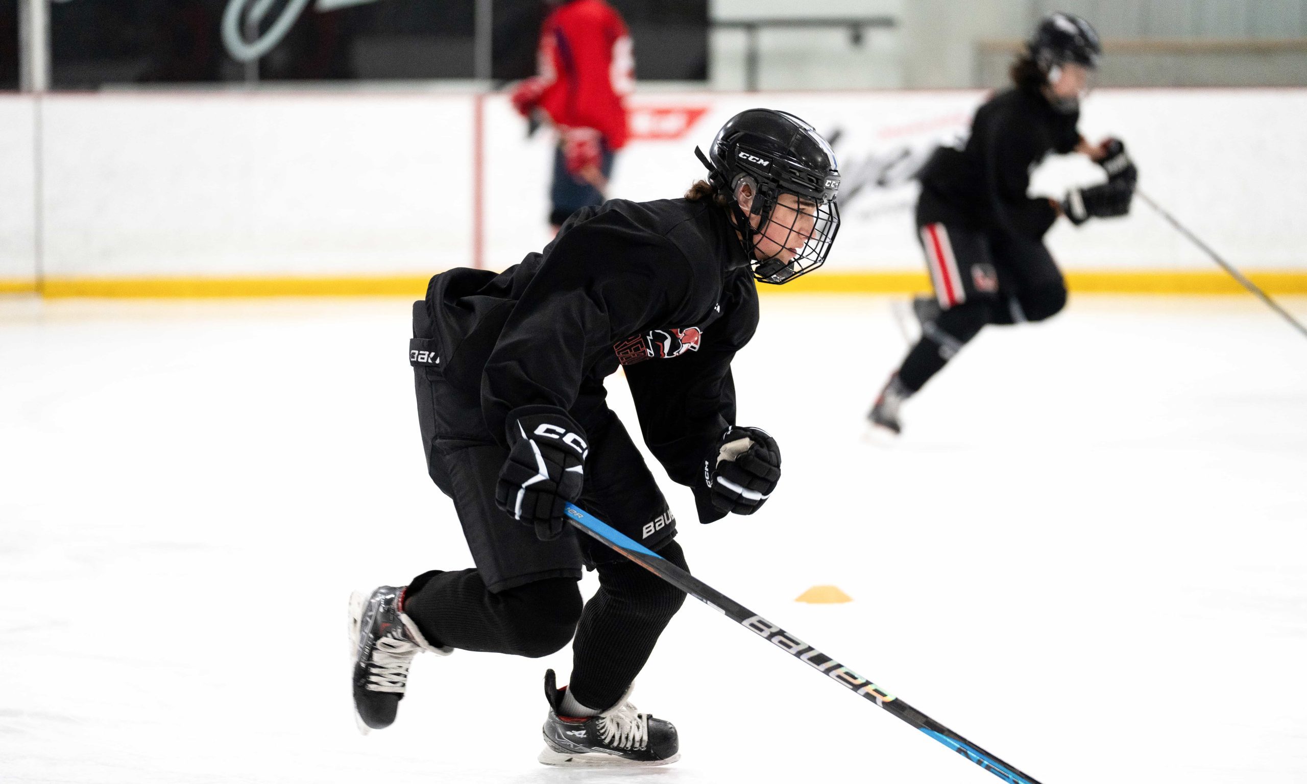 A hockey player in black gear skates on an ice rink. The player holds a stick, with teammates in the background, conveying energy and focus.