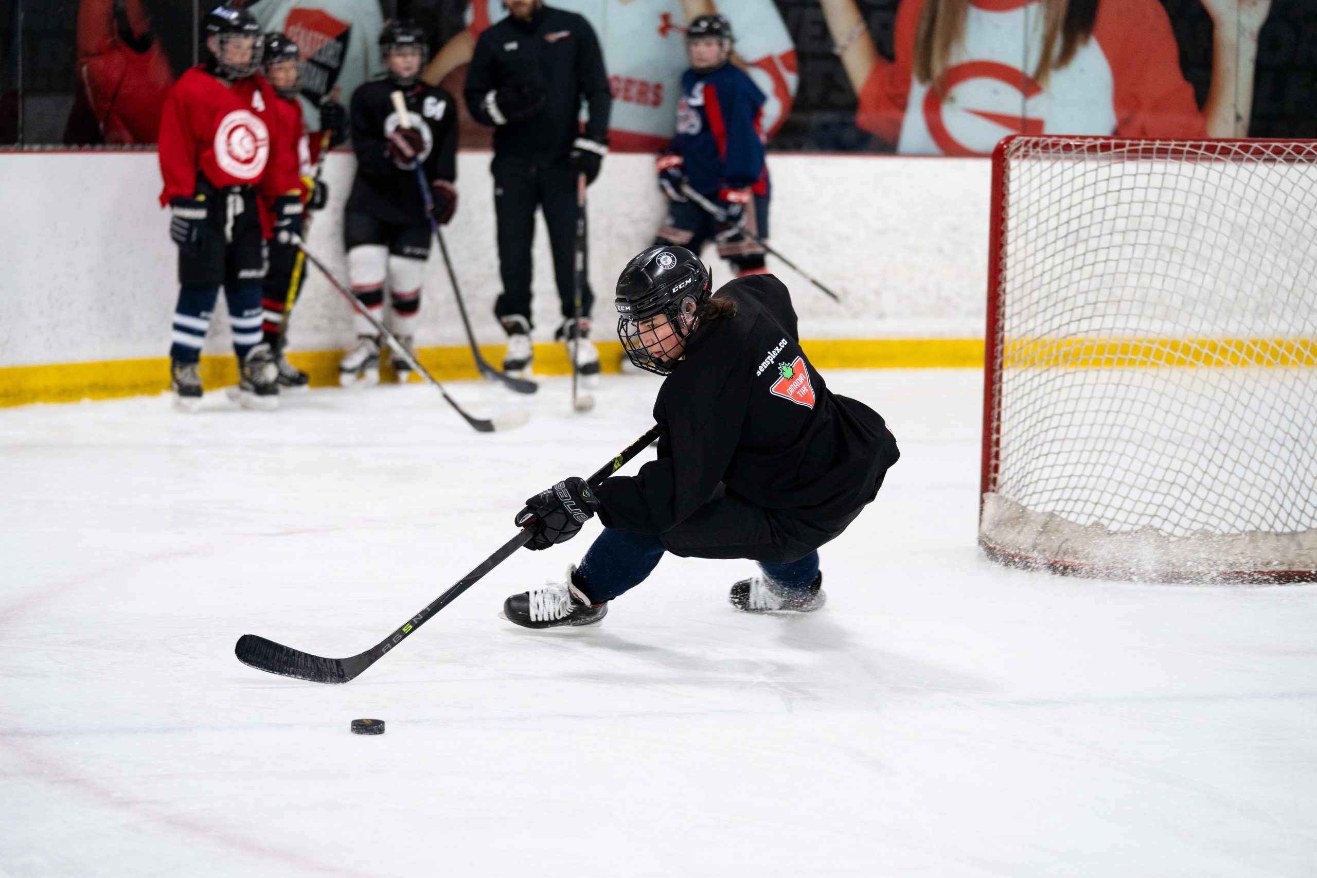 Hockey player in black jersey taking a shot on ice as other players watch near the goal.