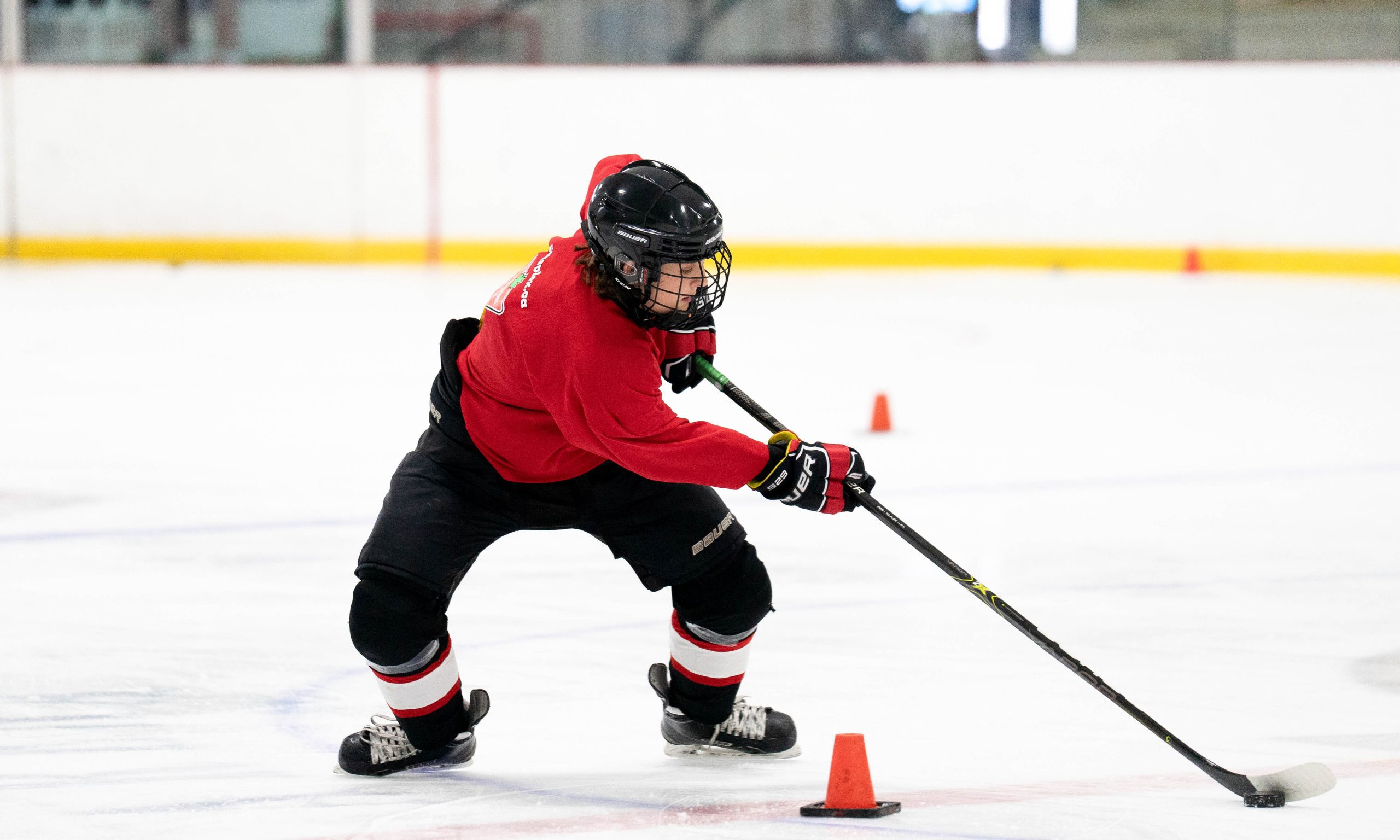 Hockey player in a red jersey and black pants skillfully maneuvers a puck around cones on an ice rink, showcasing agility and focus.