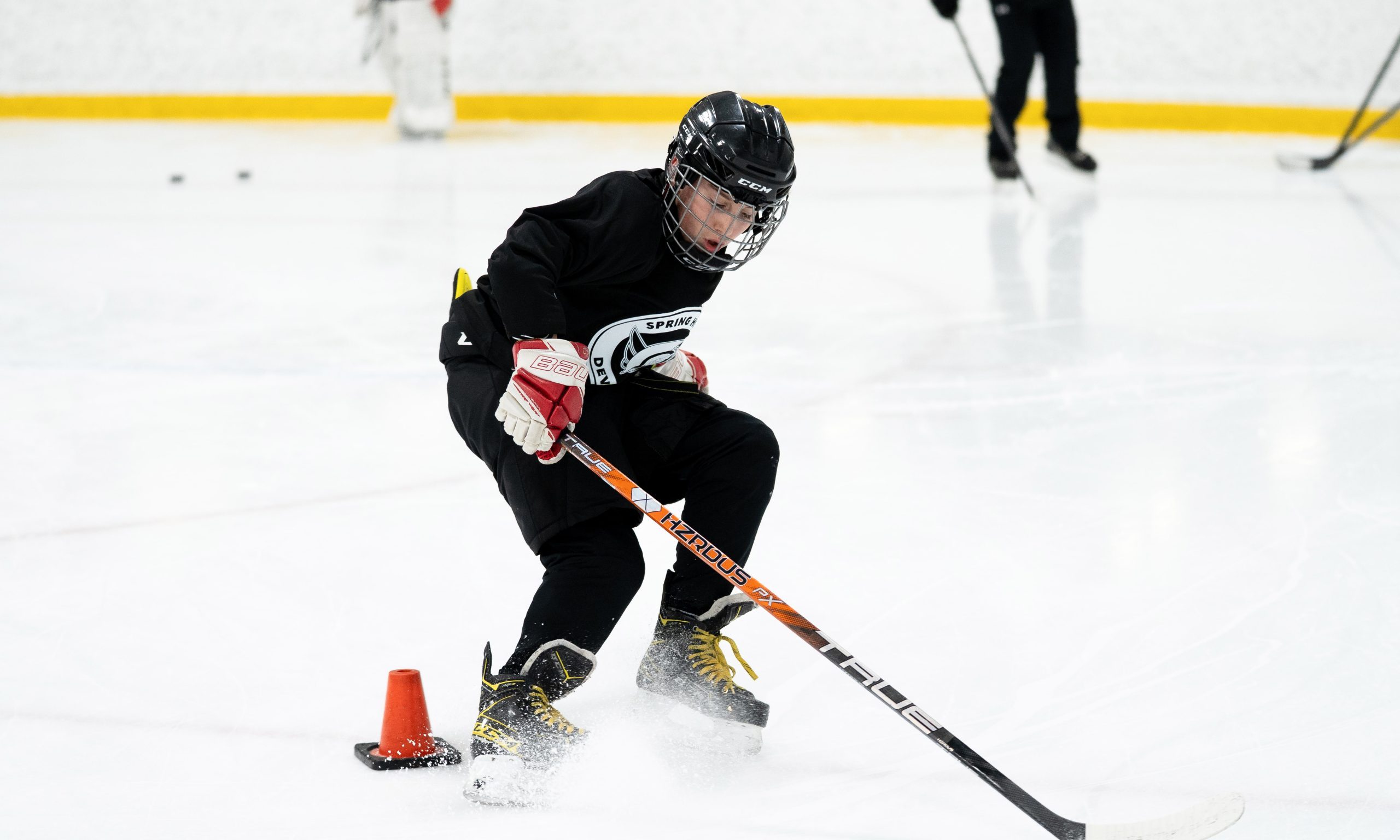 A young hockey player in black gear skillfully pivots around an orange cone on ice.