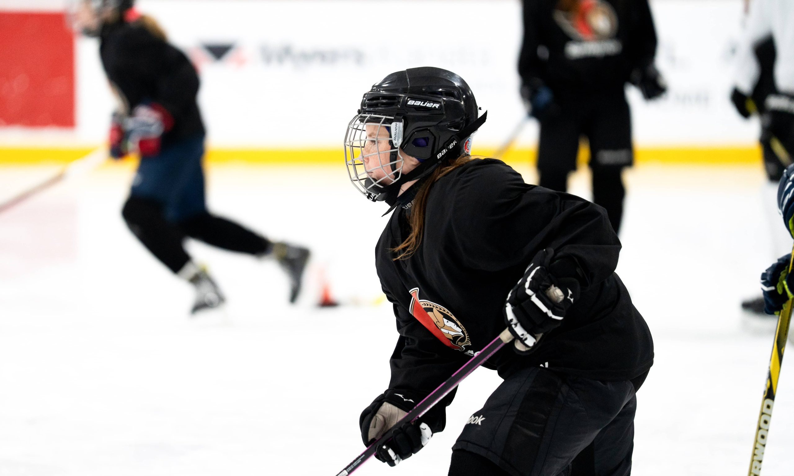 A young girl in black hockey equipment skates on an ice rink, focused and determined.