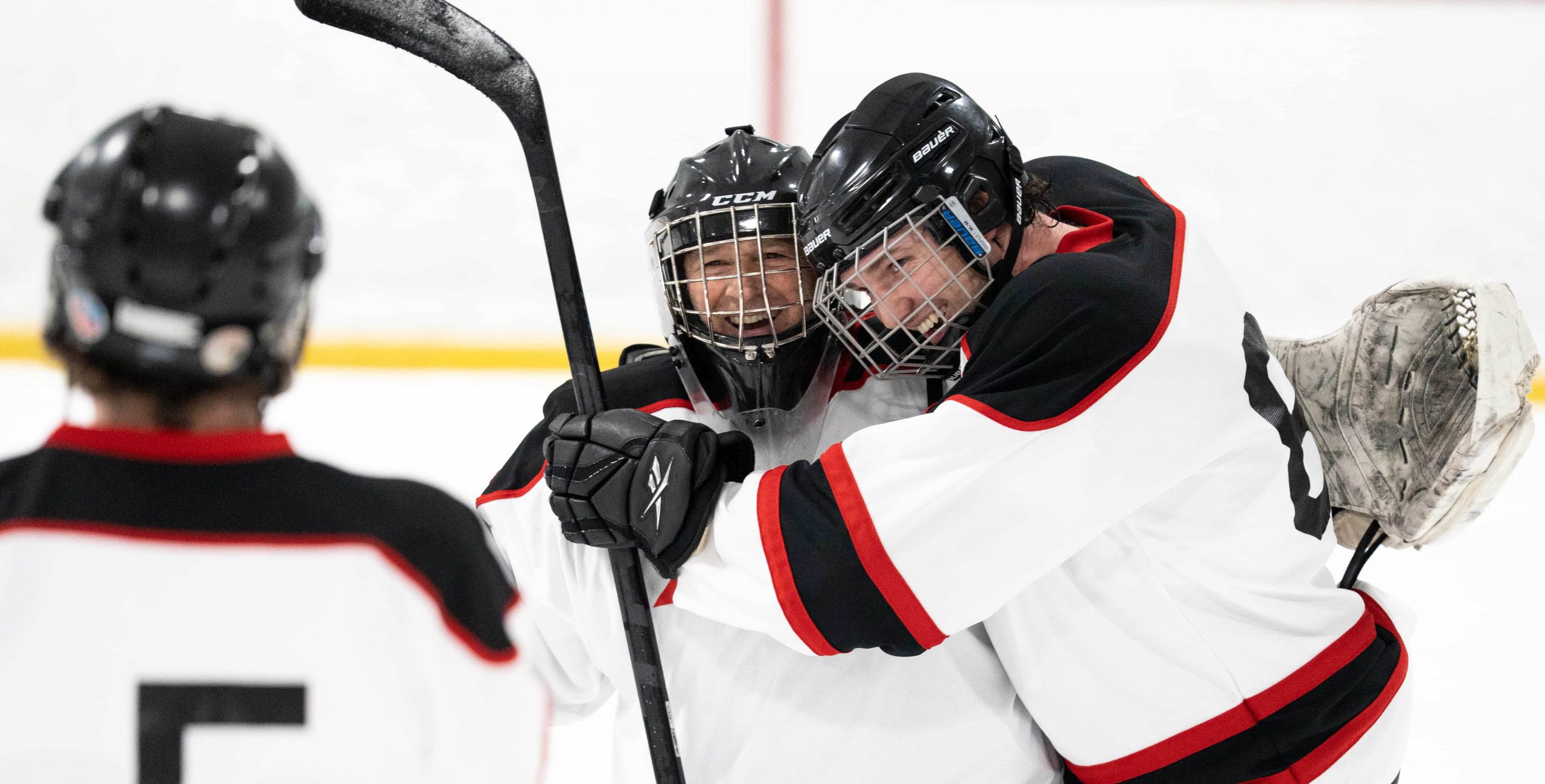 Two hockey players in white jerseys hug on the ice while a third teammate stands nearby, partially visible.