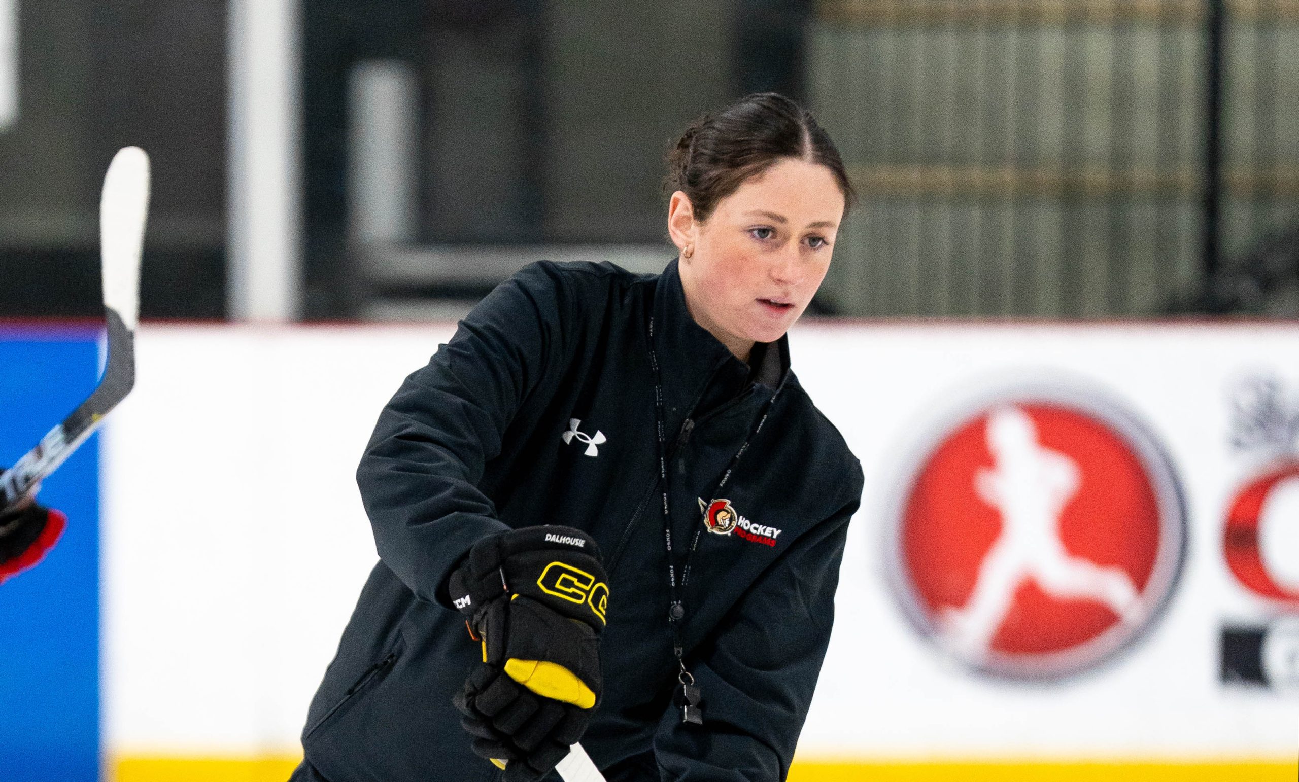 A hockey coach in a black jacket and gloves holds a hockey stick on an ice rink, focused and poised.