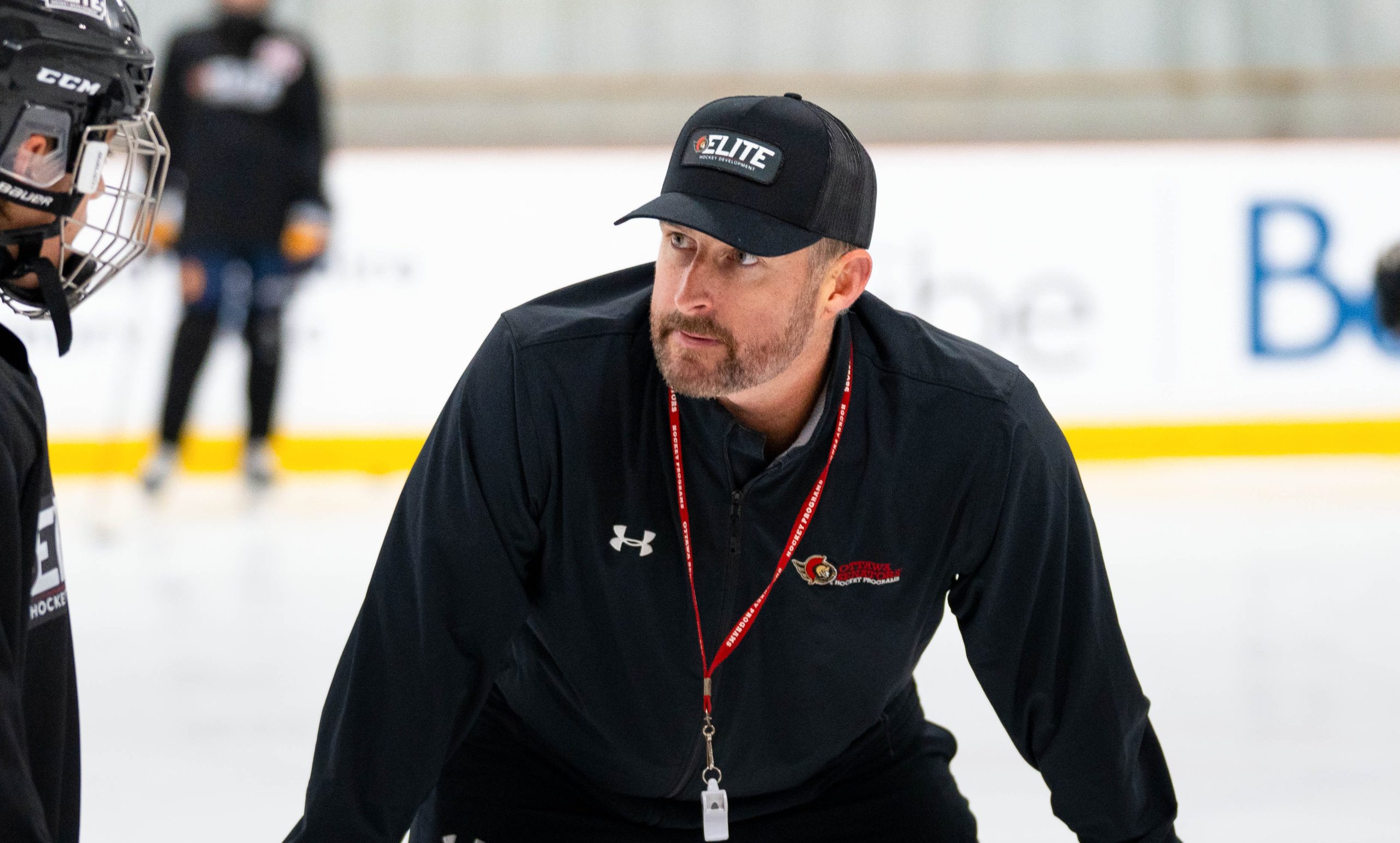 Hockey coach in black sportswear and cap talks to players on the ice during practice.