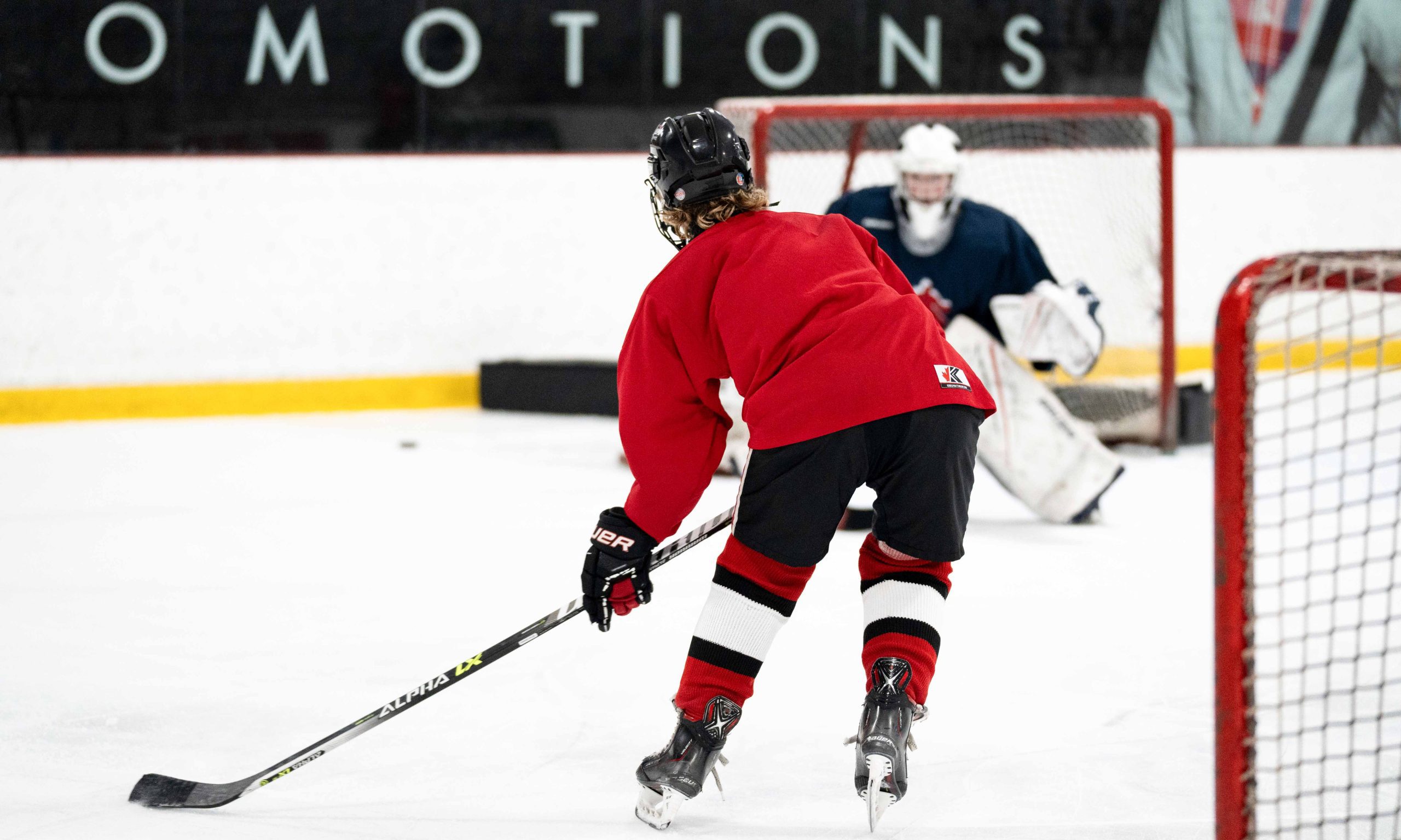 A youth ice hockey player in a red jersey skates towards the goal, holding a stick, aiming to shoot. The goalie in navy awaits, fully focused.