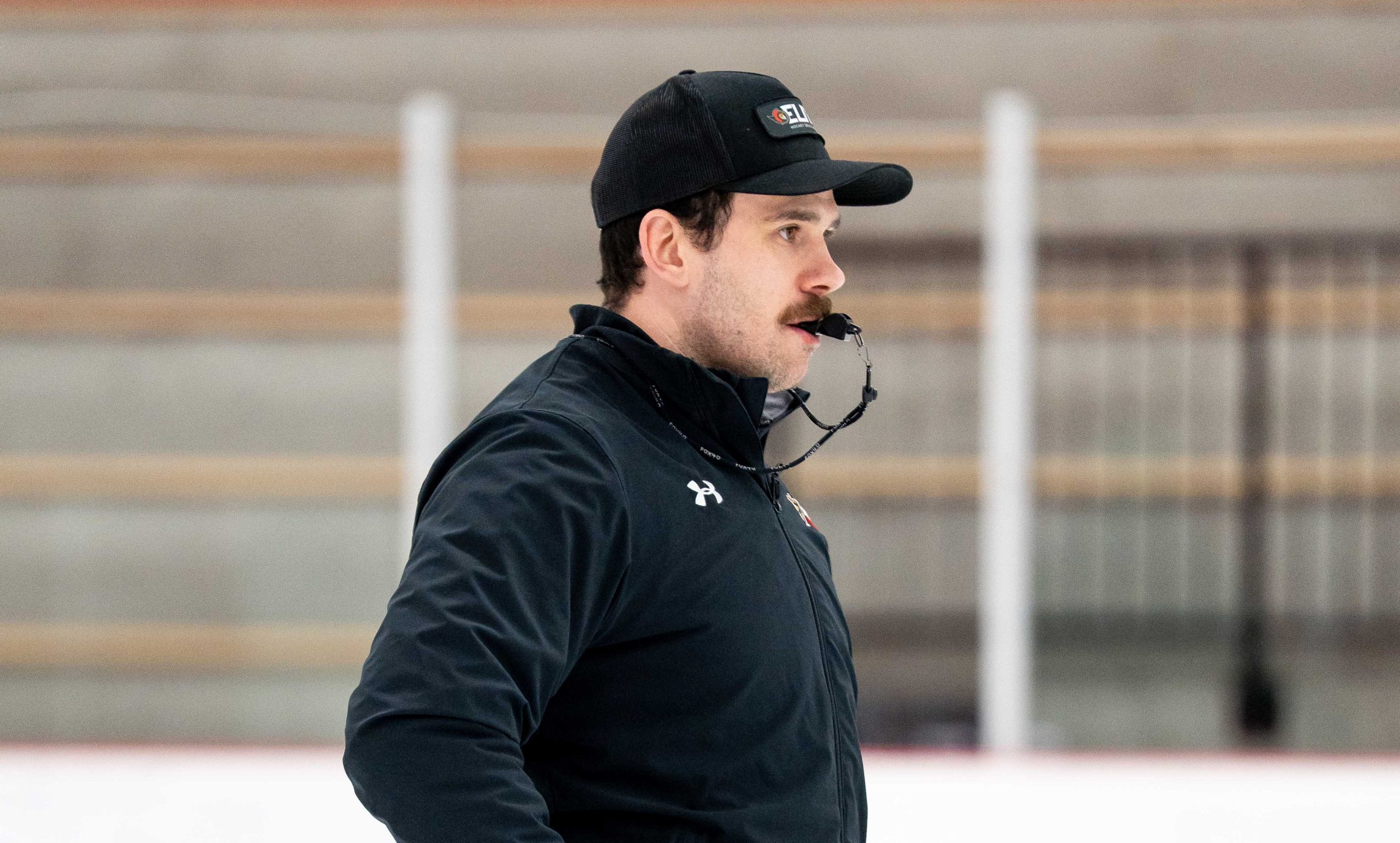 A hockey coach wearing a black jacket and cap stands on an ice rink with a whistle in his mouth.
