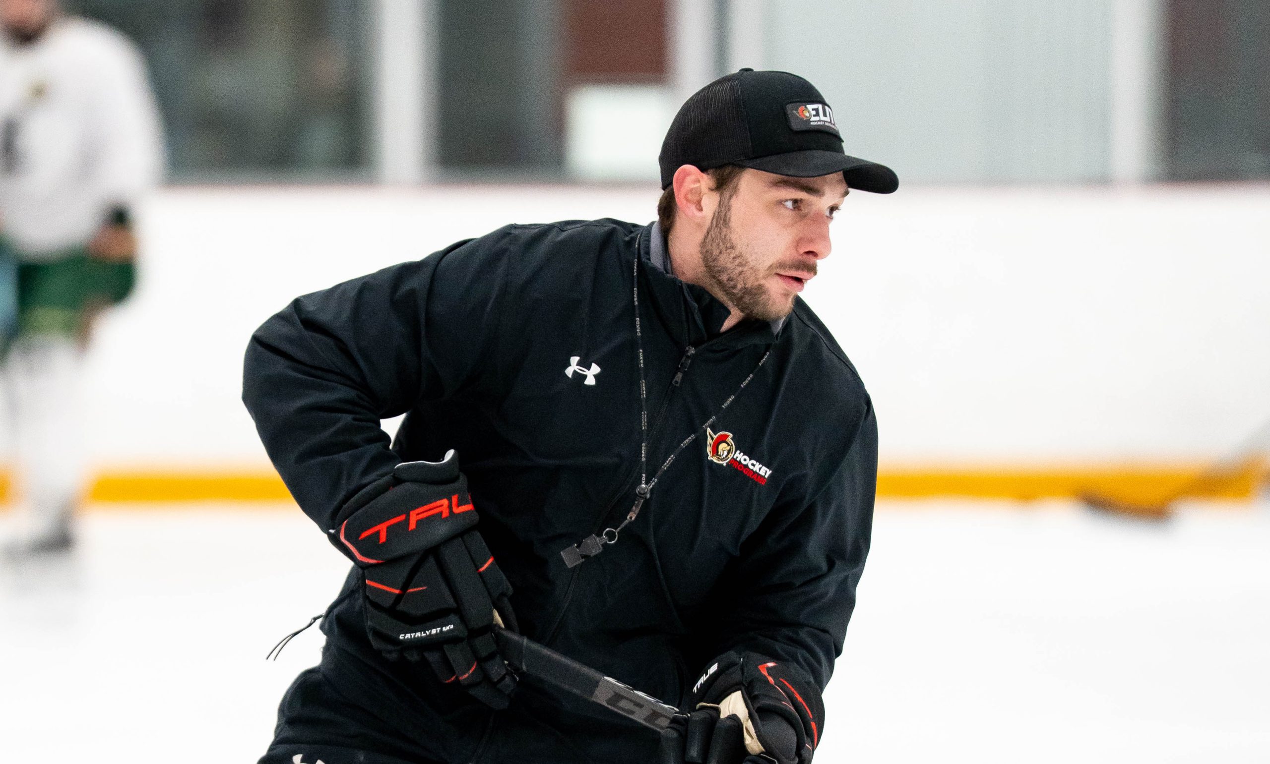 A hockey coach wearing a black jacket and cap skates on the ice, holding a hockey stick and whistle.